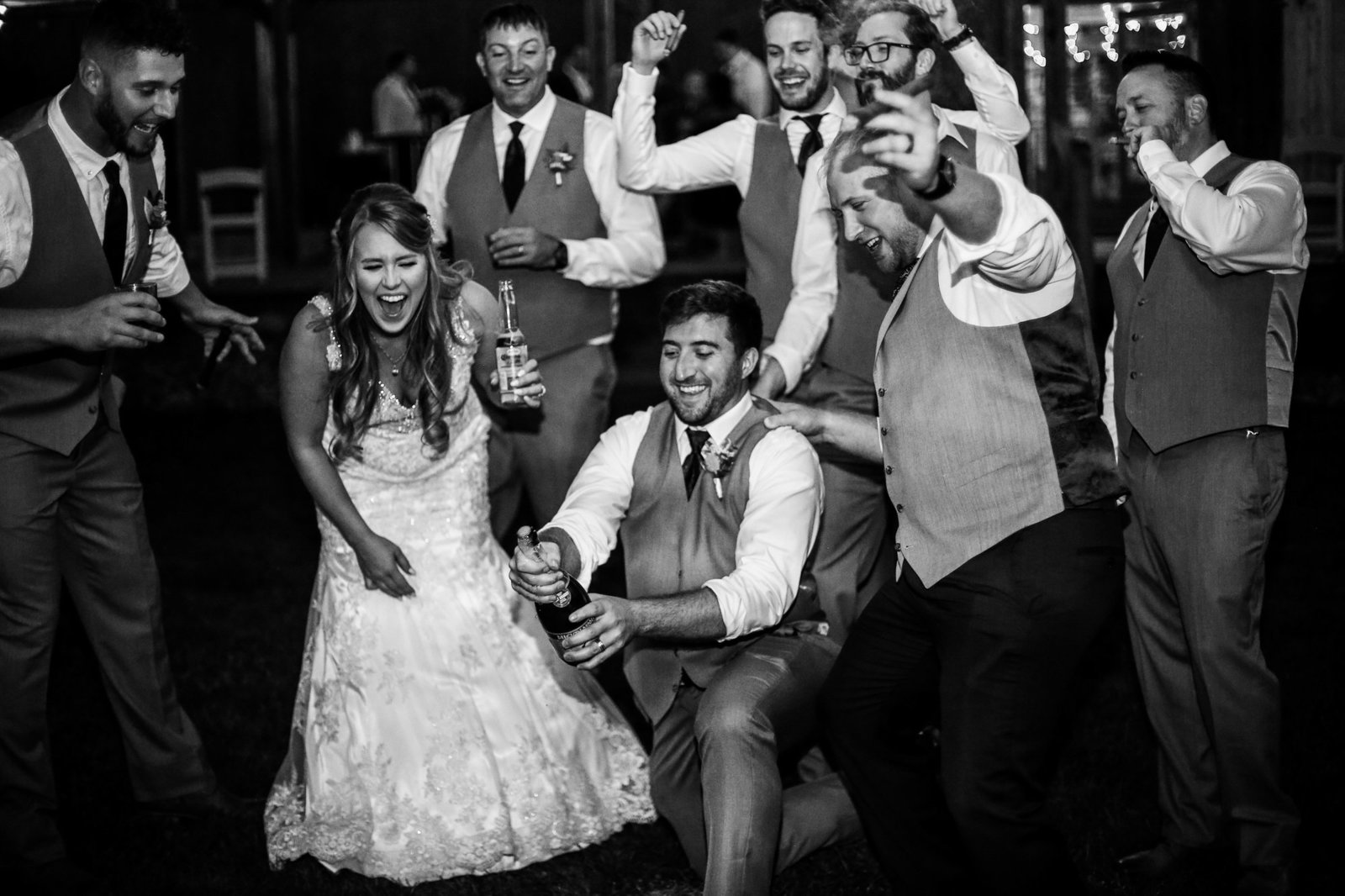 Bridal party laughs as groomsman aatempts to open a bottle of champagne