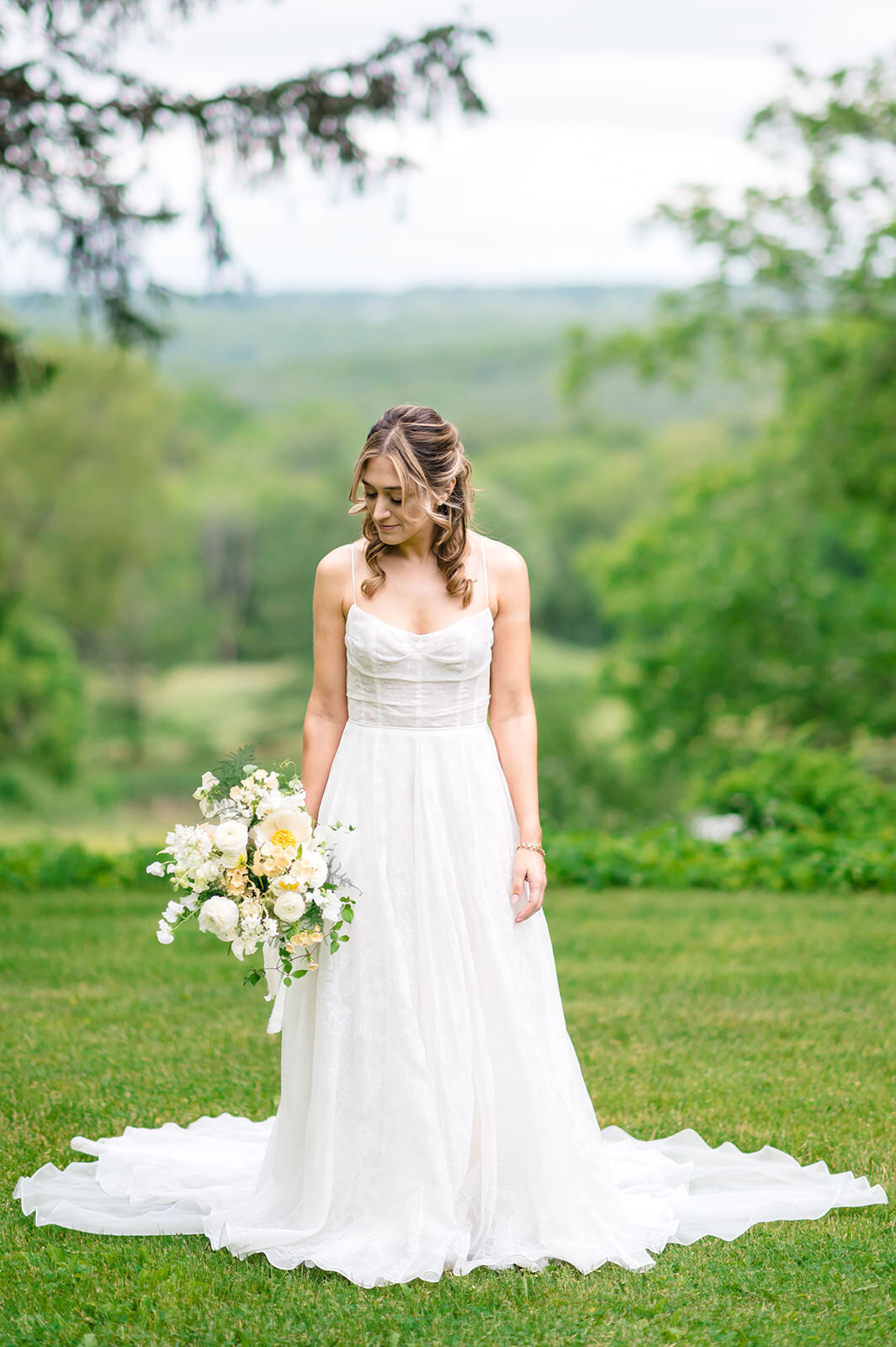 A bride in a strapless wedding dress holding a delicate bouquet stands in a lush green field with a scenic view of rolling hills in the background