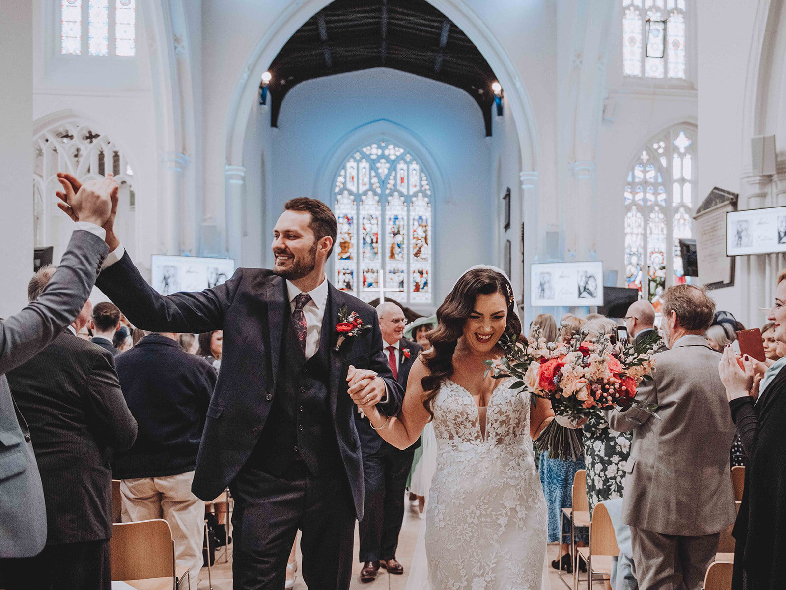 Groom exchanges a high five while bride smiles holding bouquet as they walk out of HT Cambridge having just become husband and wife