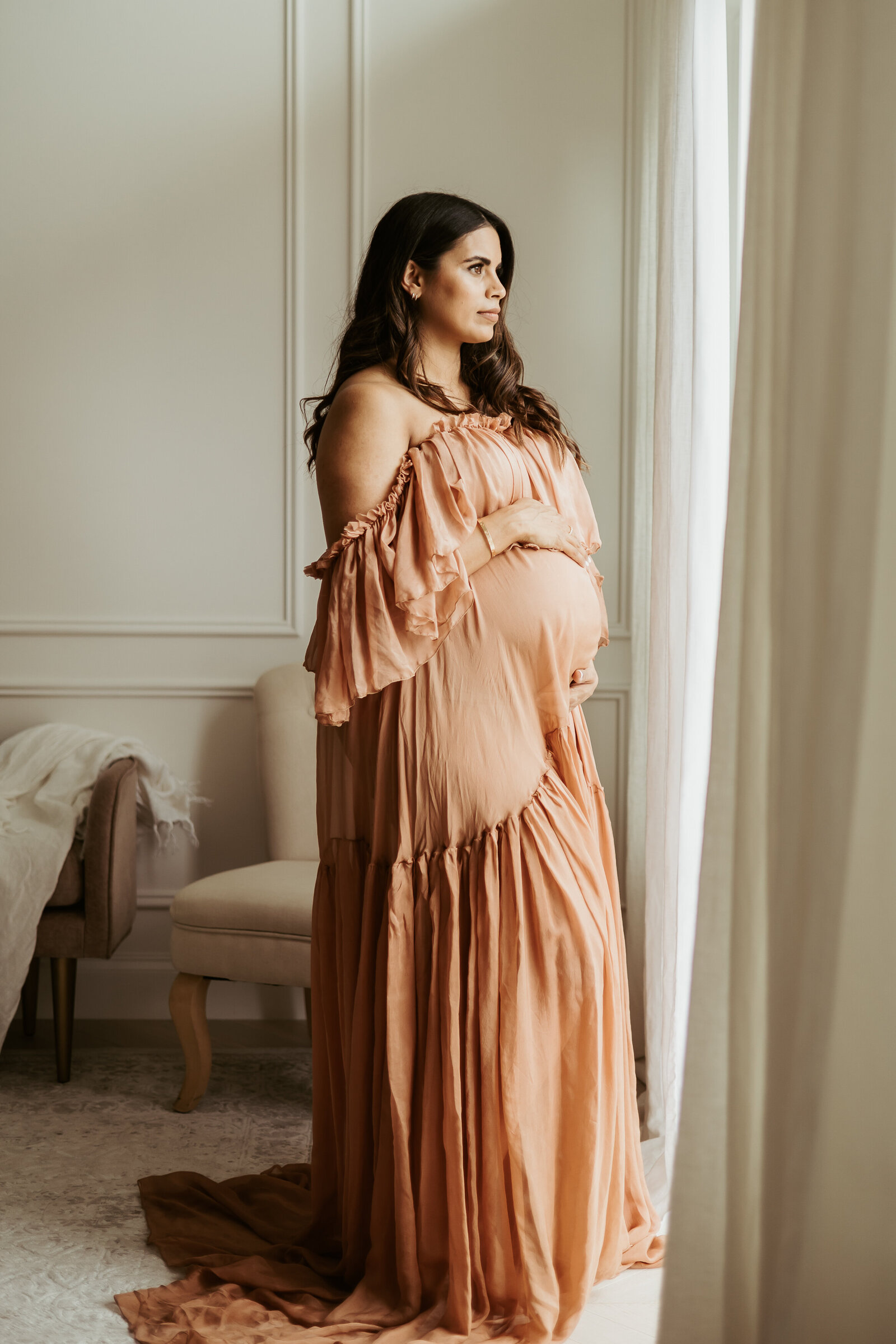Pregnant mama in a long brown silk dress cradling her bump in front of a window