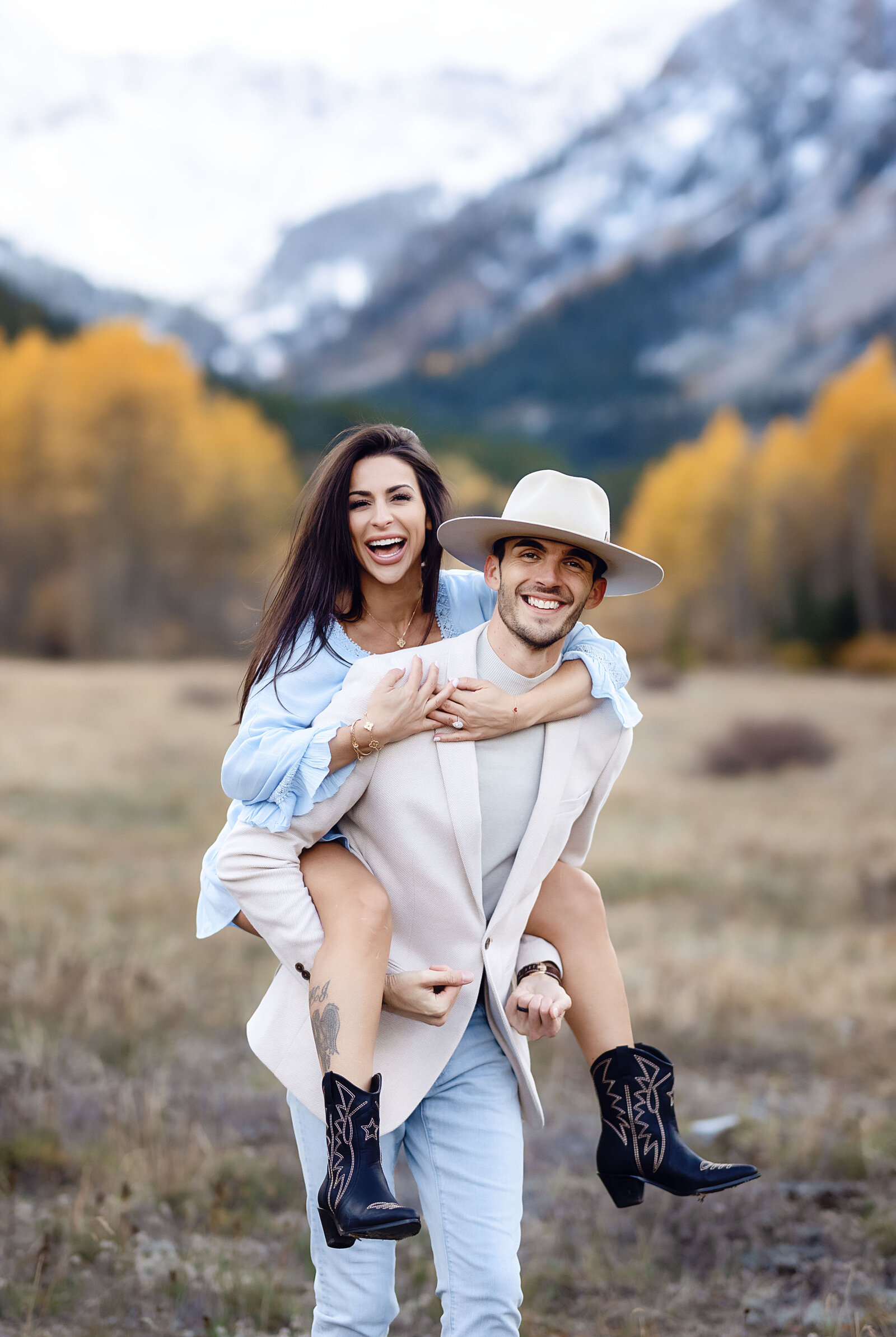A young man has his girlfriend on his back as they dance playfully in  front of the Aspen mountains.
