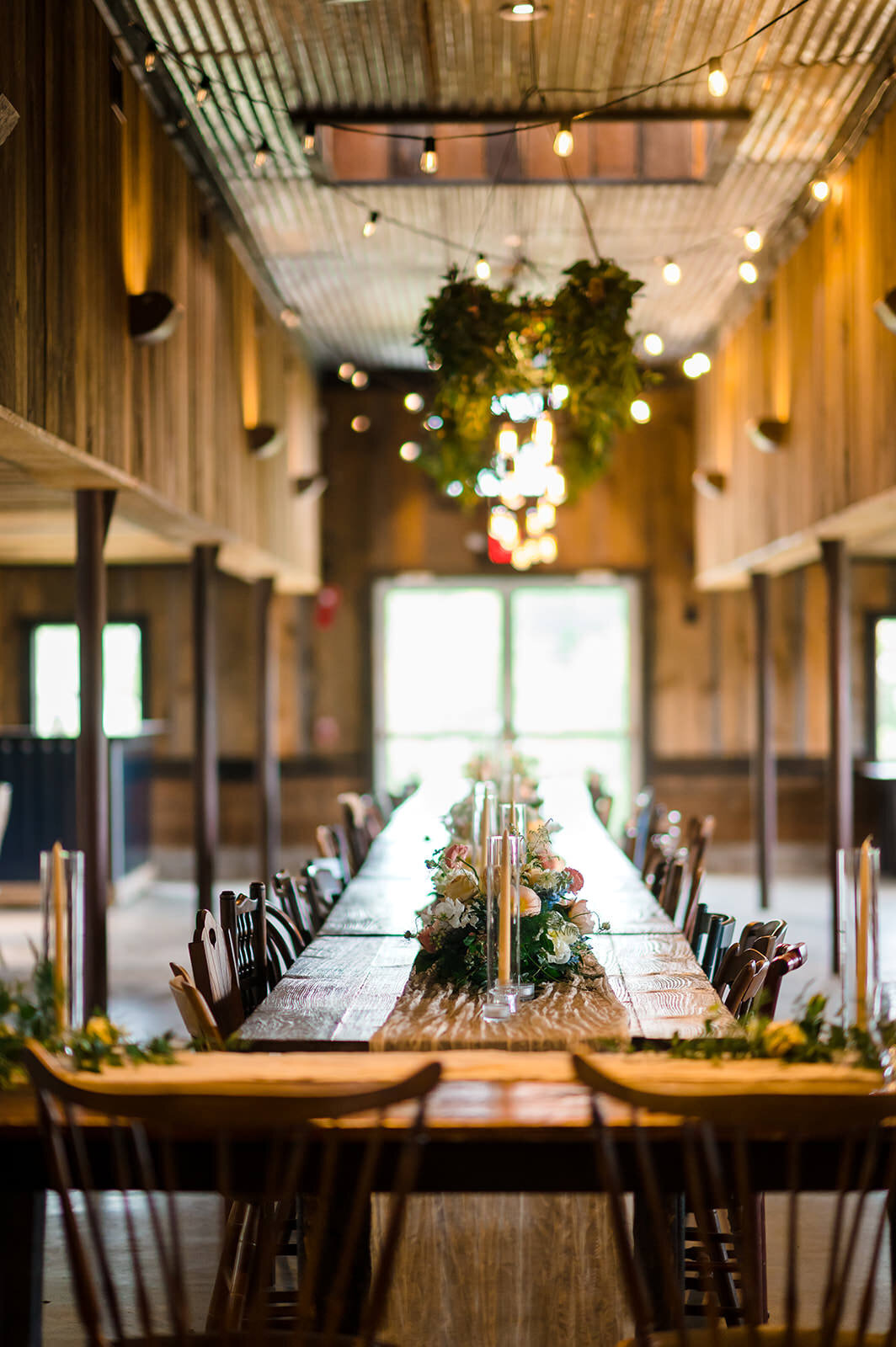 A long, elegant wedding reception table set in a rustic barn venue, adorned with natural greenery, flowers, and warm, ambient lighting