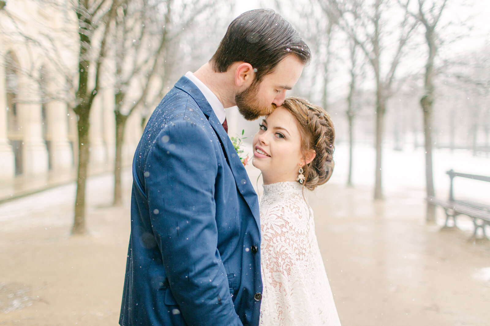 bride looking at camera as the groom kisses her forehead and snow falls around them