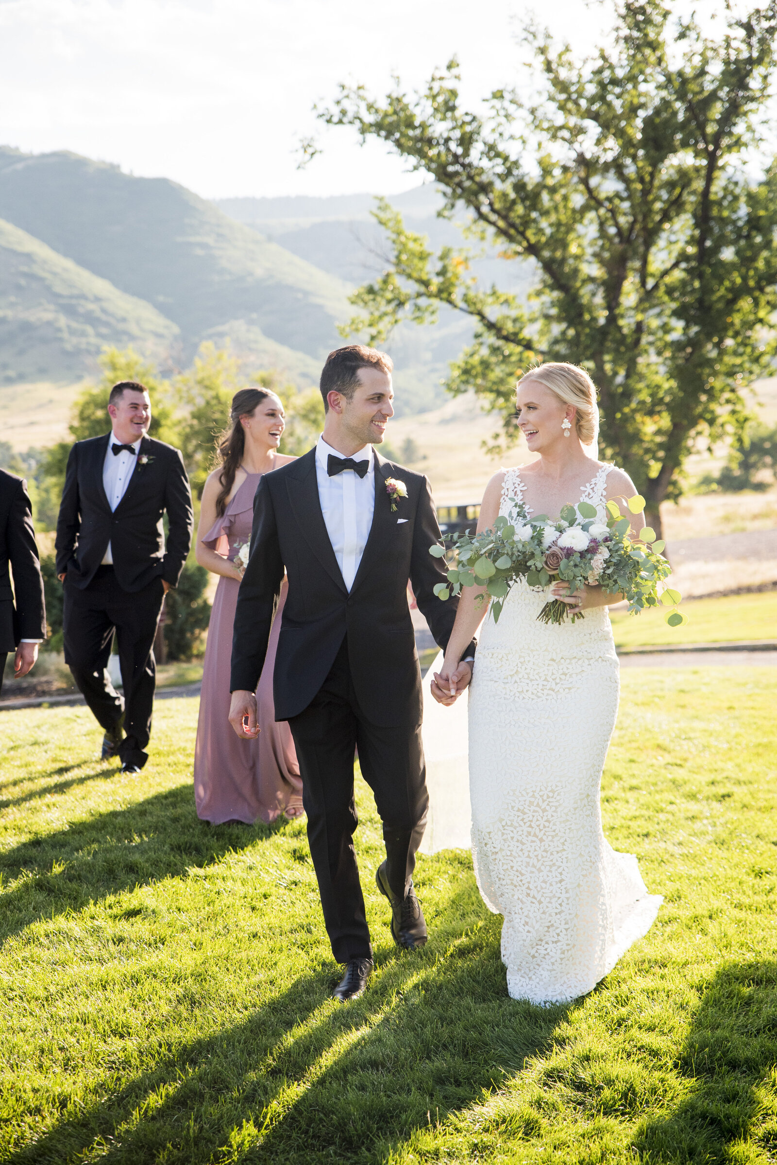 A bride and groom walk hand-in-hand toward the camera with their wedding party in the background, captured by Littleton wedding photographer Two One Photography.