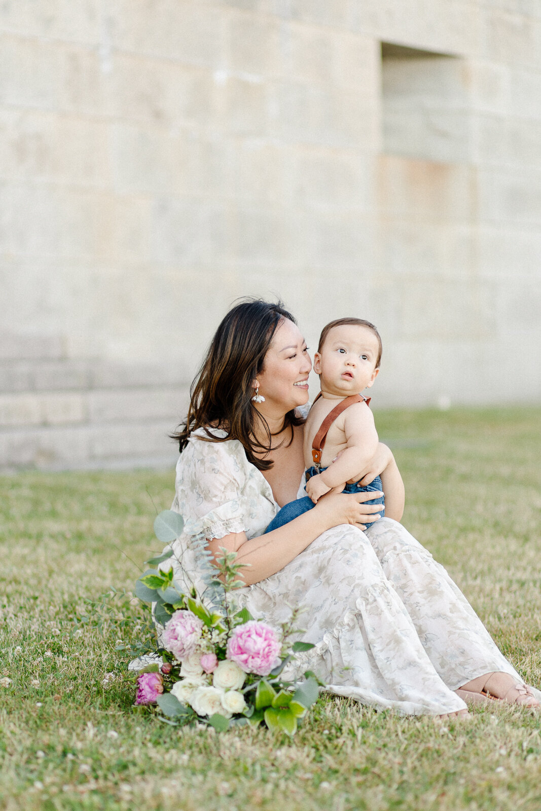 A mother wearing a floral gown sitting on the grass with a floral arrangement placed in front of them,  holds her baby boy during photo session with Boston photographer Corinne Isabelle