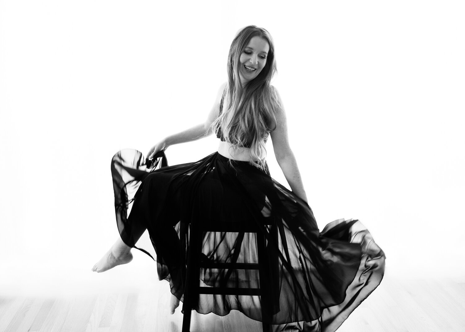 Black and white image of a woman in a San Francisco boudoir studio, caught in a moment of carefree motion. She's wearing a black flowing skirt that flares out as she twirls, paired with a delicate black top. Her long hair sways with the movement, her smile radiating happiness.