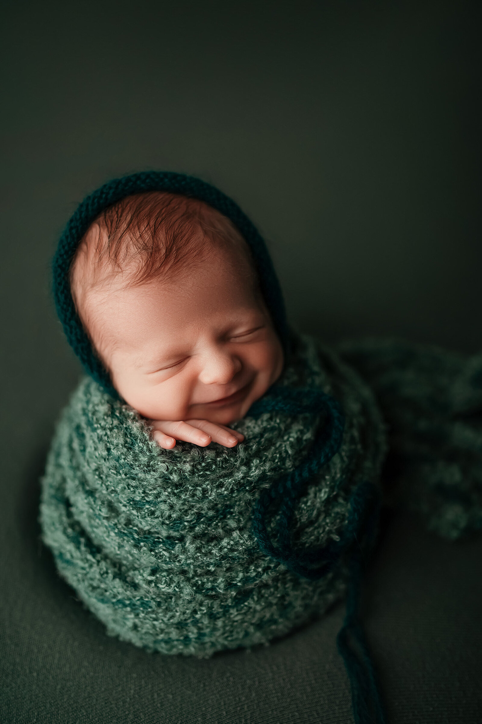 A smiling newborn is wrapped in green fabric and styled with coordinating green hat and backdrop in our cozy Waukesha studio.