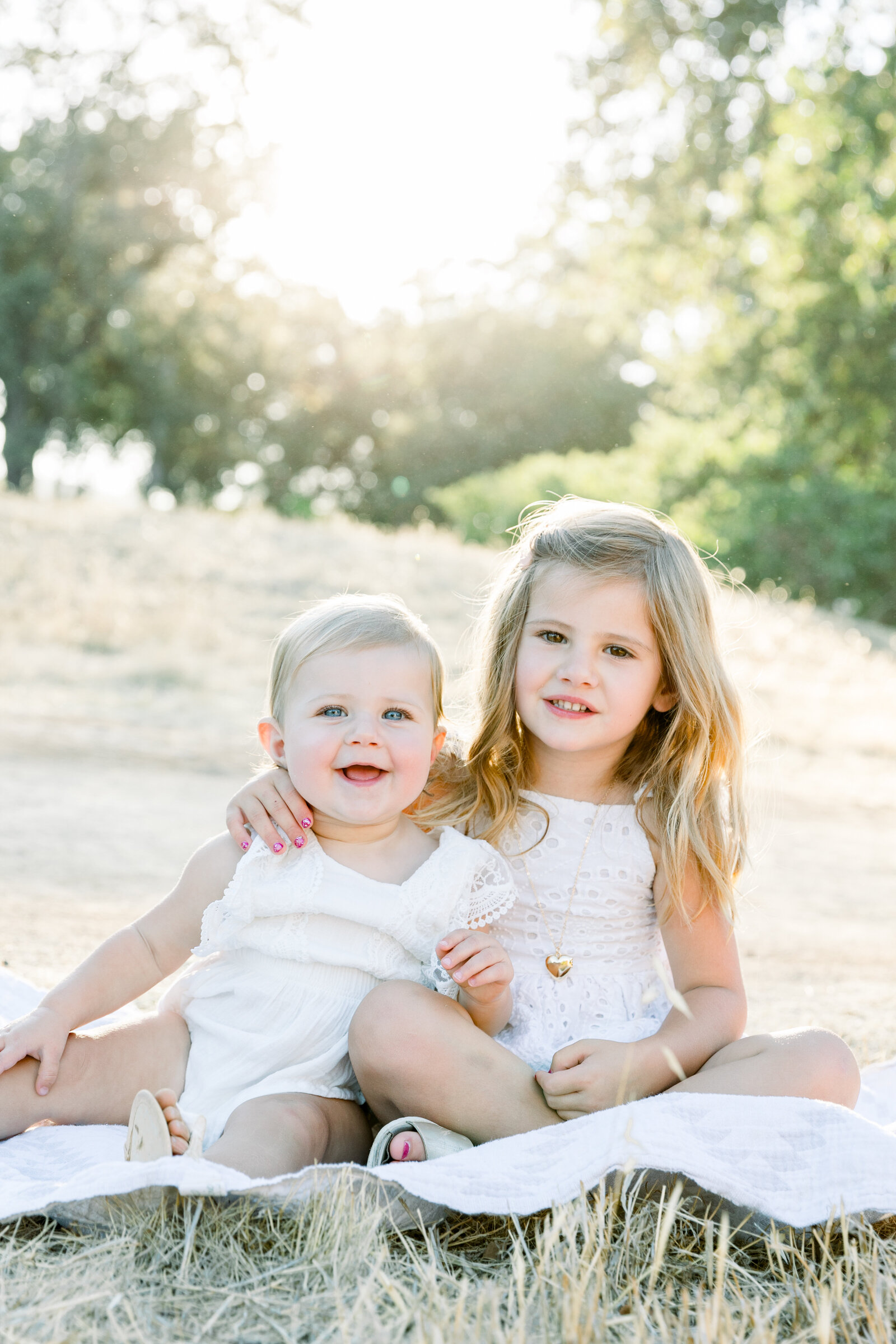 Image of two sisters sitting on blanket in a field together smiling taken by Sacramento Newborn Photographer Kelsey Krall