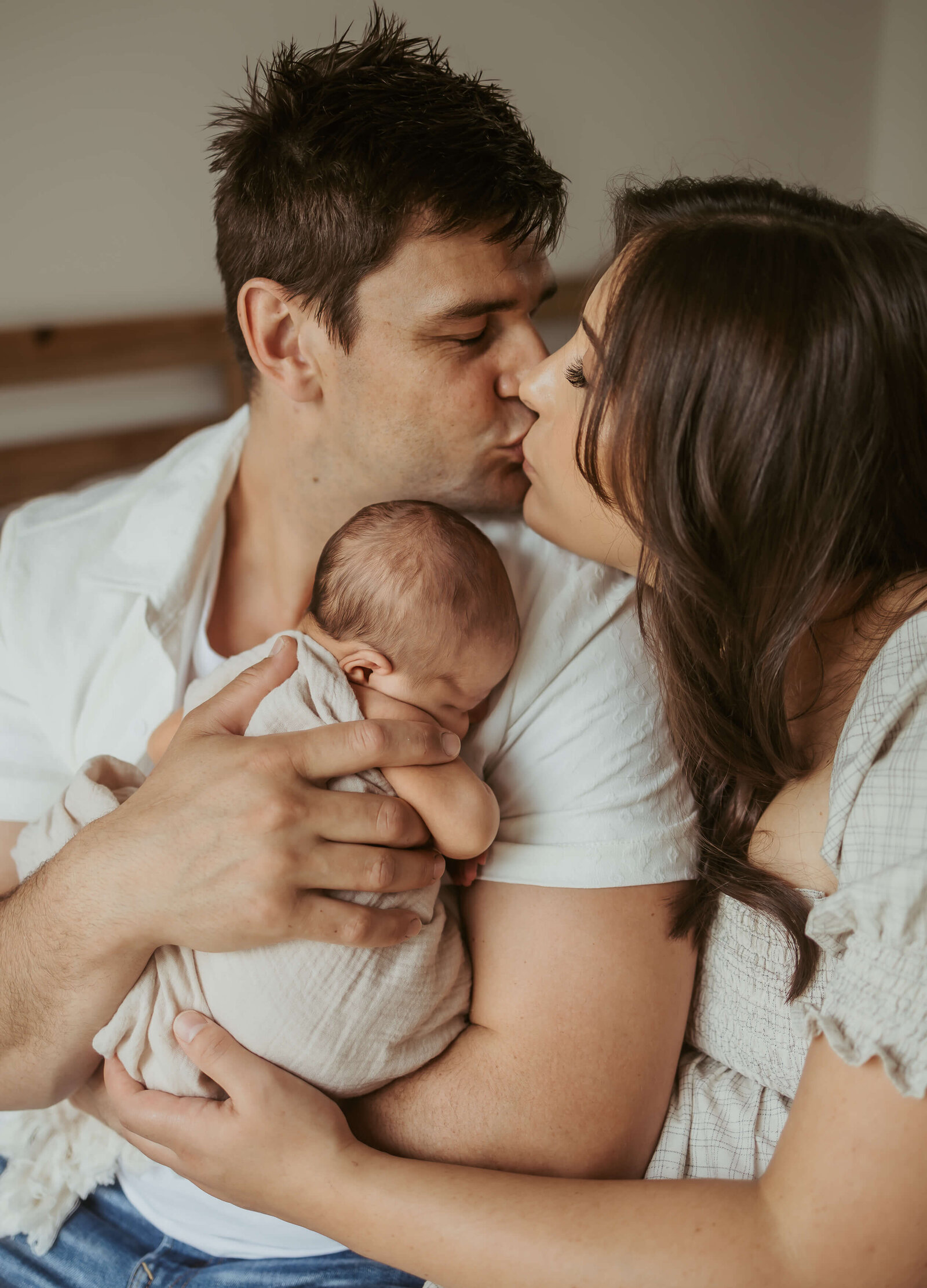 New parents are kissing while dad holds his new son upright on his chest while he's sleeping.