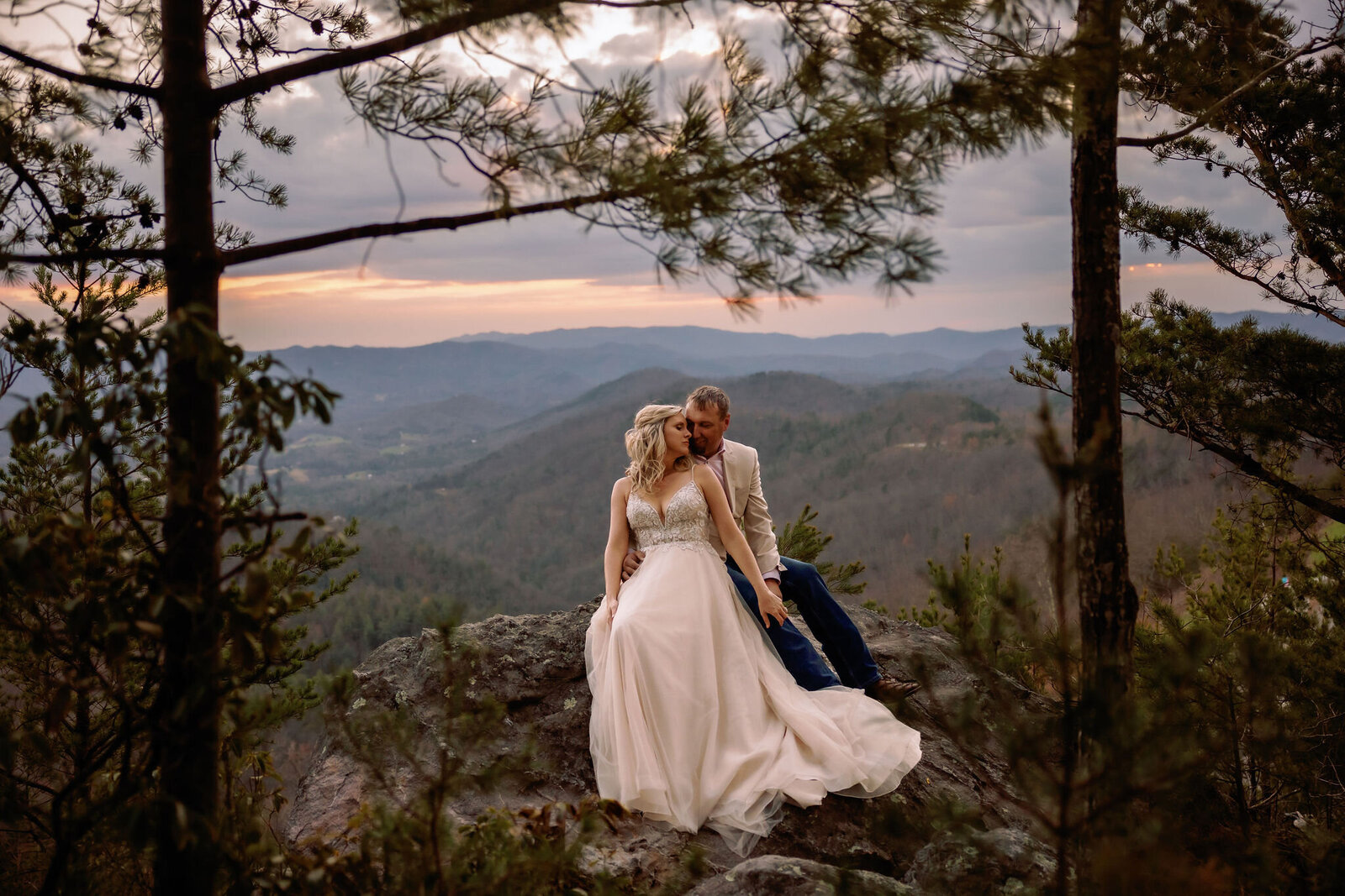 foothills wedding pictures captured by gatlinburg photographers of couple eloping to Gatlinburg and sitting on a large boulder with the Smoky Mountains in the distance as the couple cuddles and kisses 