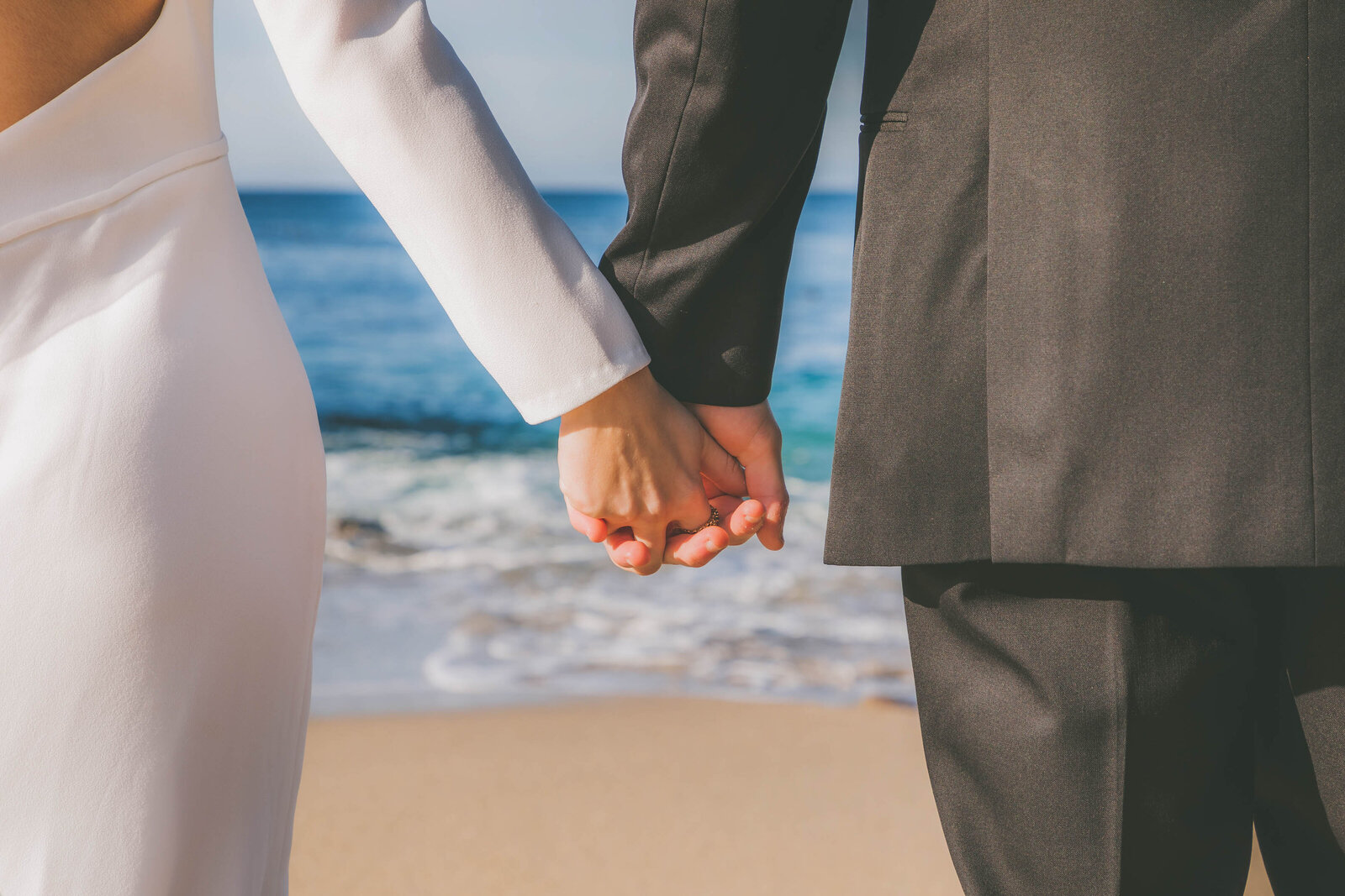 A closeup photo of a bride and groom holding hands at the beach.