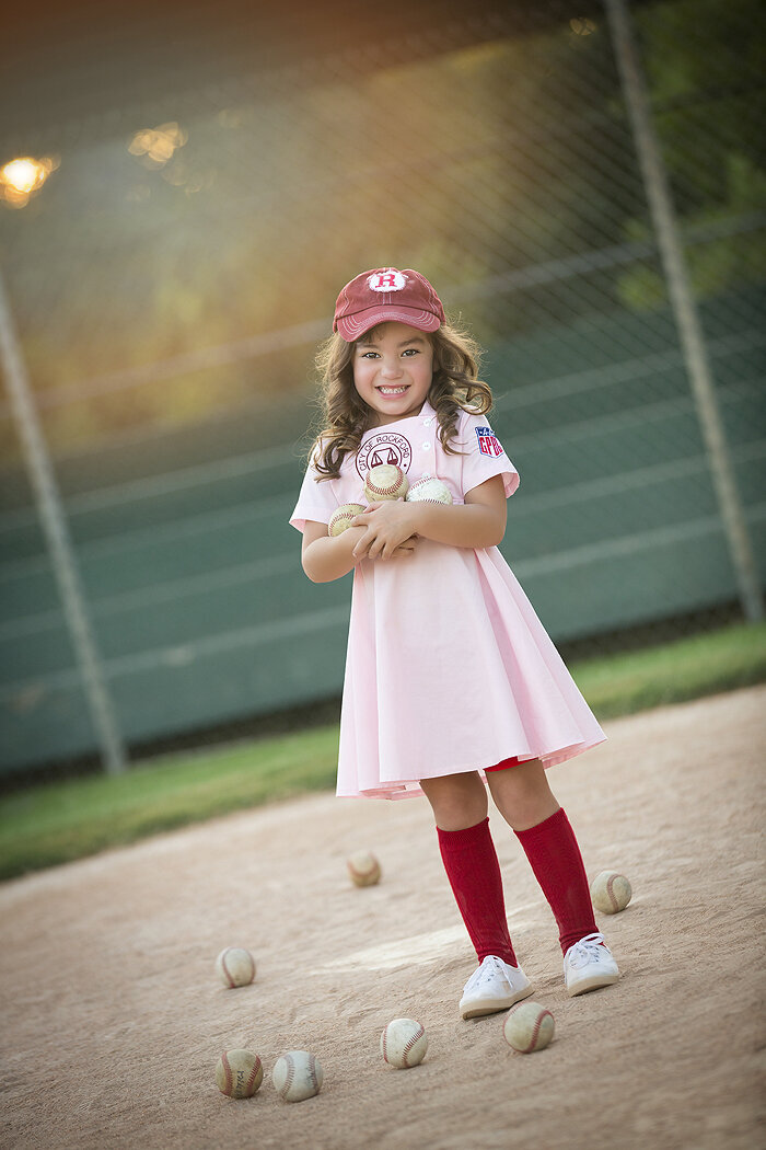 Little girl on baseball field in pink League of Their Own dress, Dallas child photography.