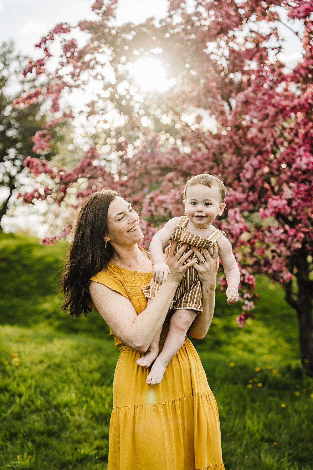 mom holds baby boy in overalls up in air at sunset with pink flowering trees