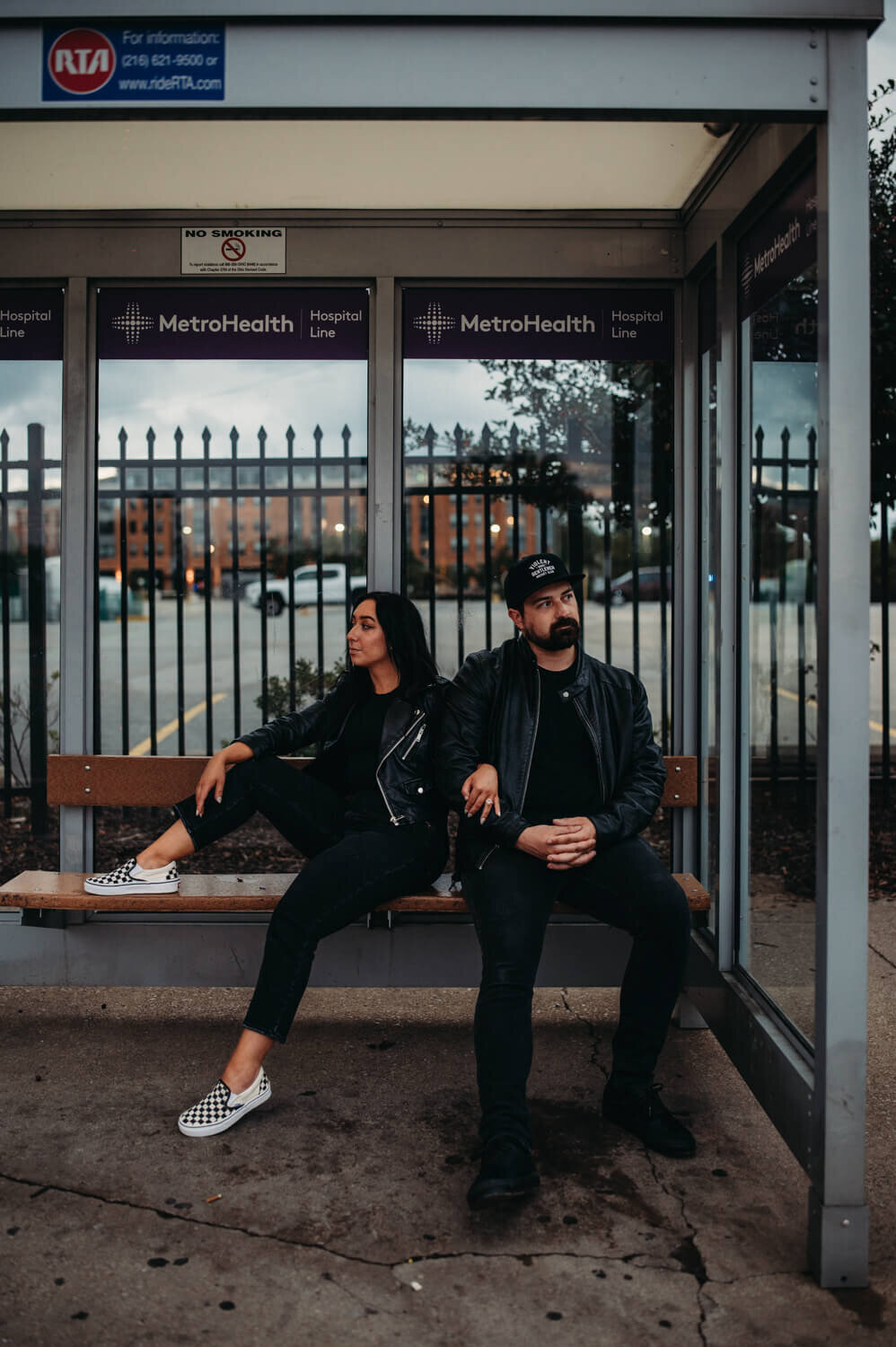 Couple sit at bus station in alternative clothing.