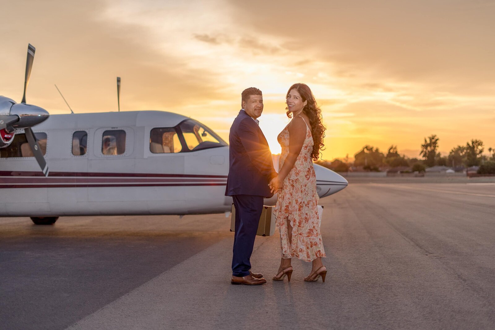 Hispanic couple holding suitcases looking back before walking around the front of plane with sun setting.