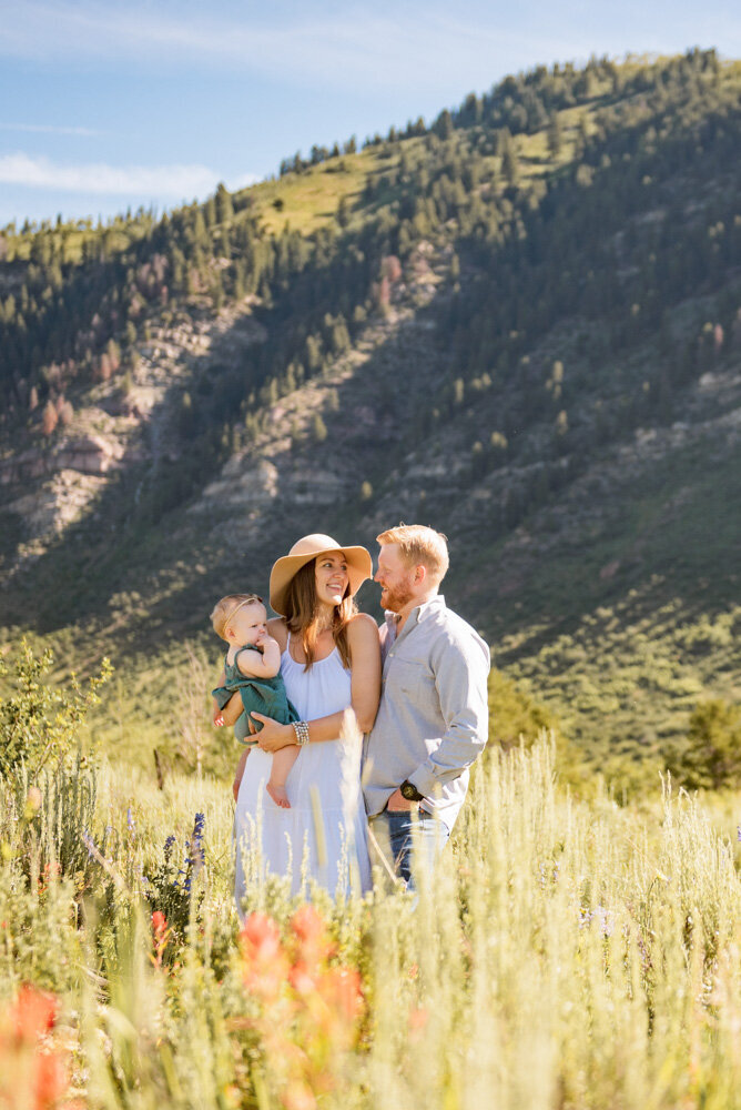 a mother in a white dress looks at her husband and holds her baby in a mountain meadow