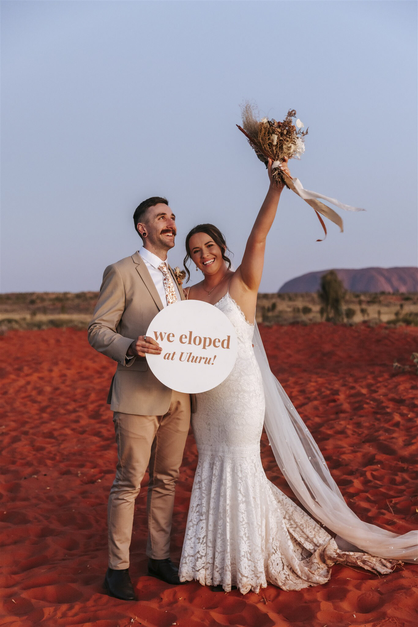 A bride and a groom holding a sign that says We Eloped at Uluru