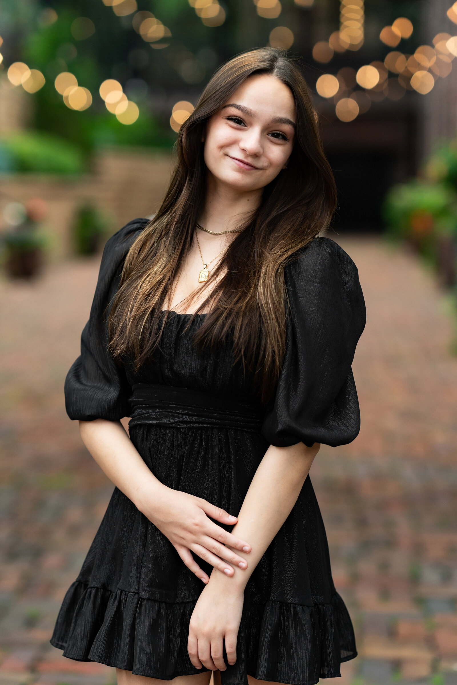 Senior girl in black dress smiles while standing on cobblestone.