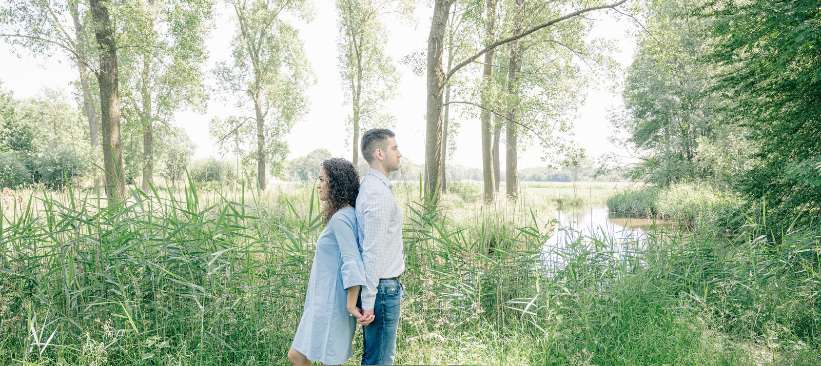 Fiancees Naomi and John stand back to back by the river Dommel in their engagement shoot