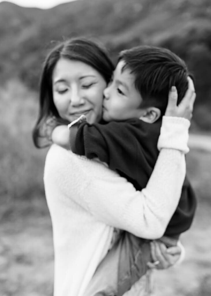 Black and white photo of mommy tightly holding her son while he kisses her cheek.