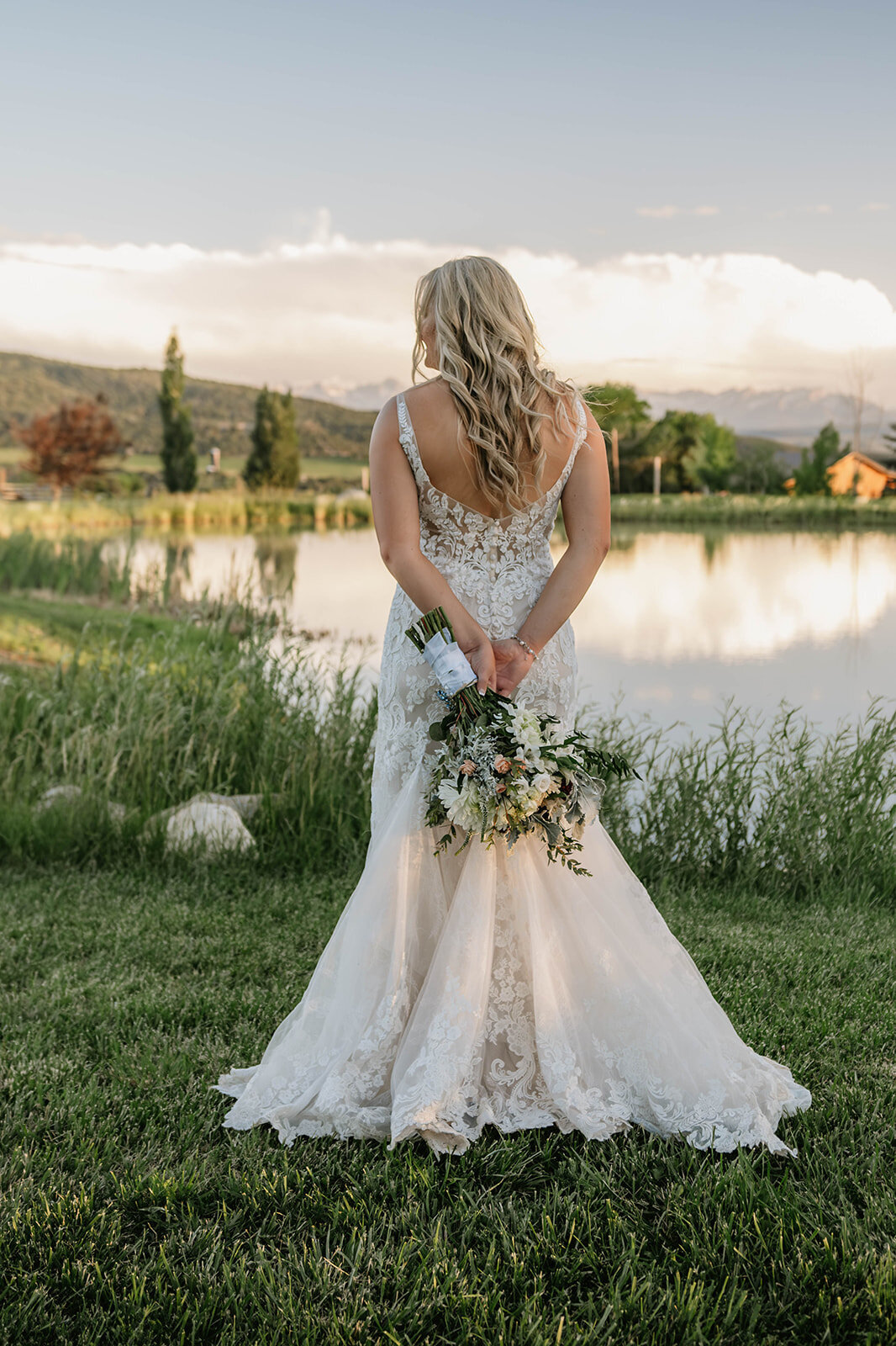 Bride in lace wedding dress stands by lake, holding bouquet behind her, with trees and mountains in the background—an adventure elopement with emotional vows captured by a Colorado elopement photographer capturing real connection.