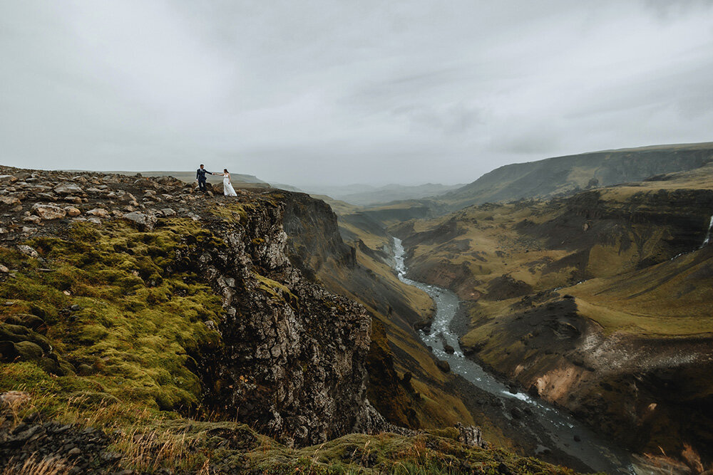 Married couple who eloped in Iceland are holding hands and standing in the edge of a cliff overlooking a green valley  in the Highlands