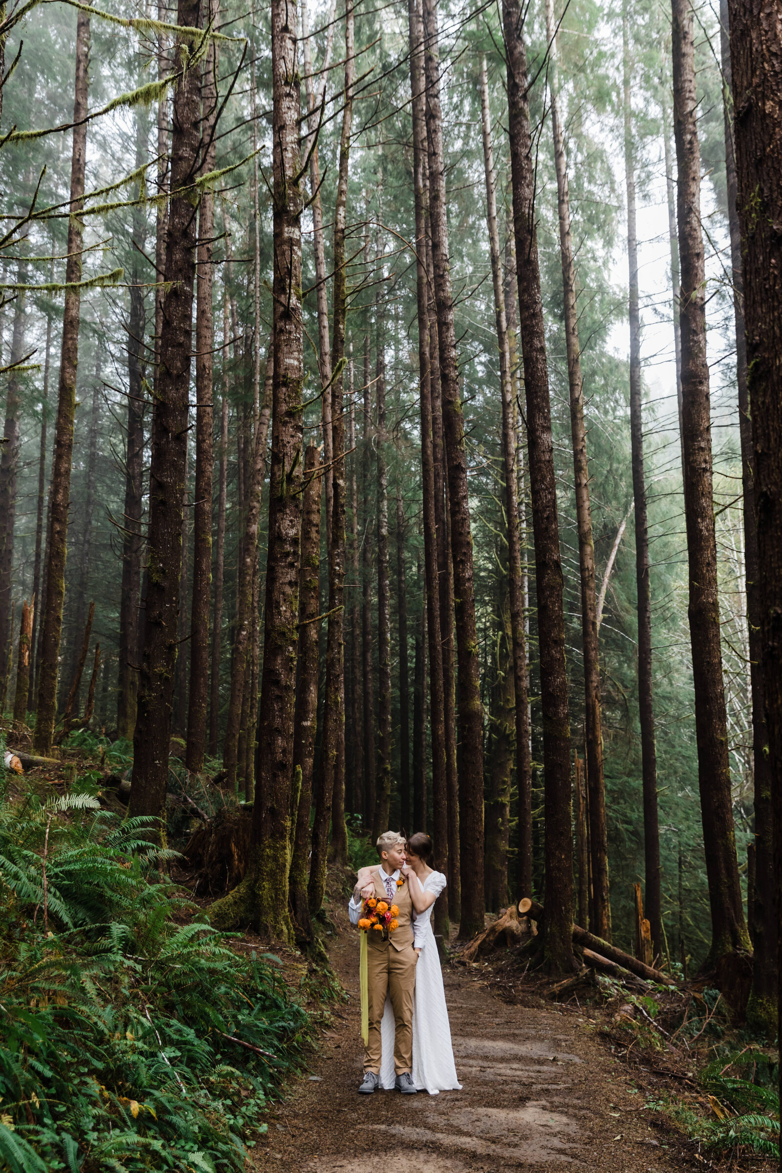 Two brides, one wearing a cap sleeve white dress nuzzled into a blond, short pixie haired bride wearing a tan vest and pants holding a bouquet of marigolds stand among the tall spruce trees for their Drift Creek Falls Oregon Adventure Elopement. | Erica Swantek Photography