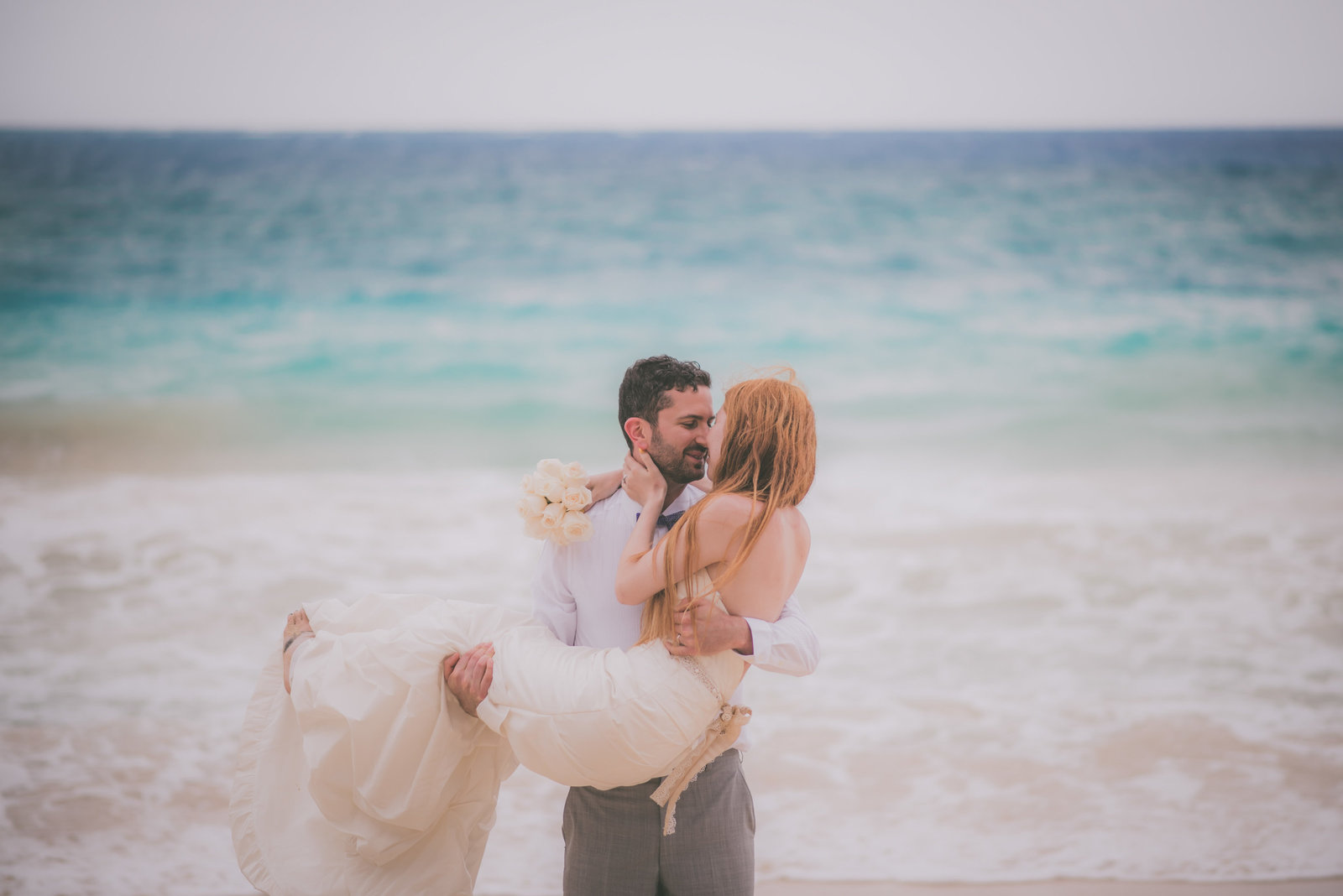 Groom holds bride on Hawaii beach while their faces get close.