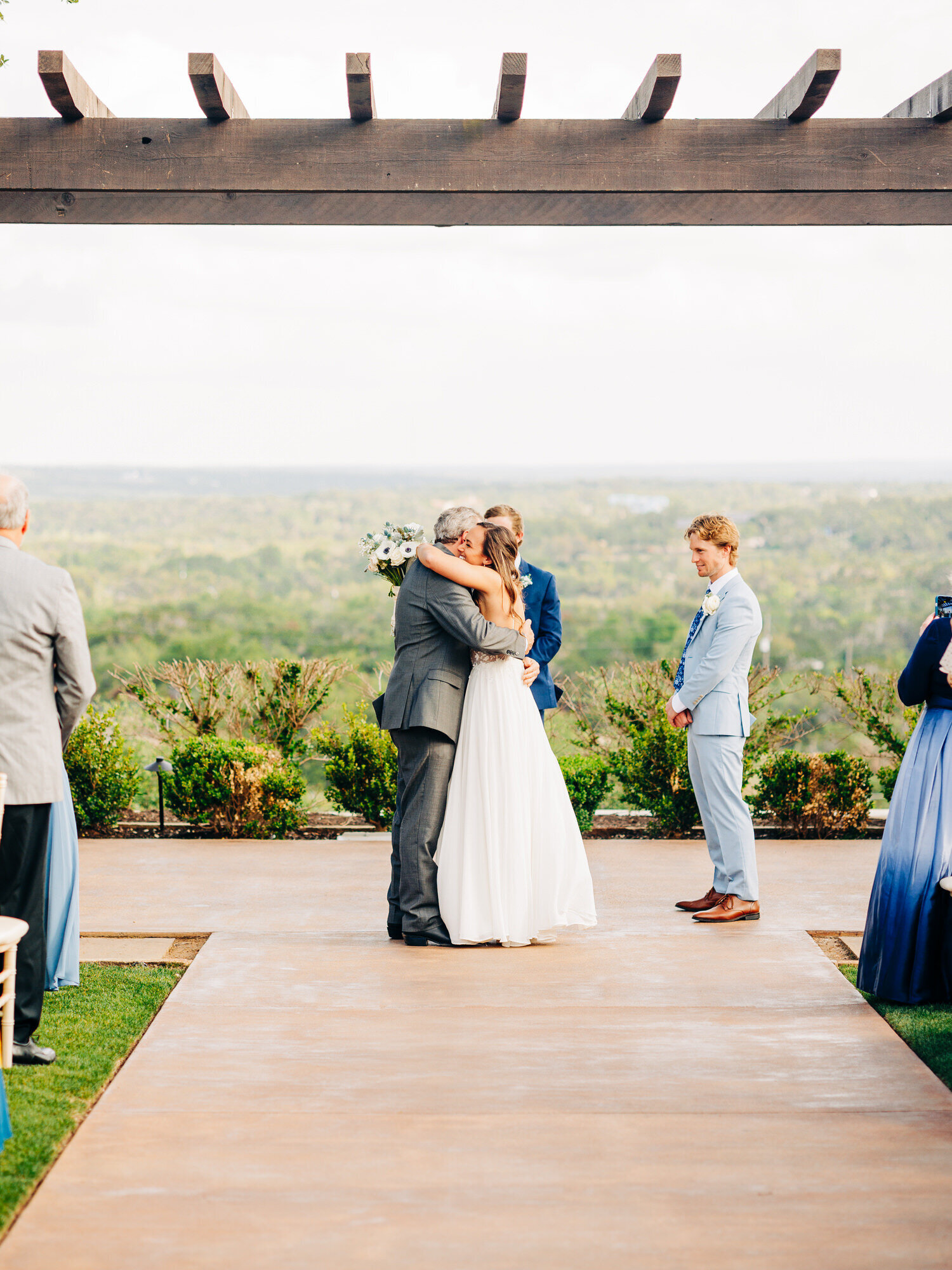 This image, taken by a San Antonio wedding photographer, features a bride hugging her dad as he gives her away at the start of their wedding ceremony. There is a view of the hill country behind them, and the groom stands to the side in a baby blue suit as he smiles.