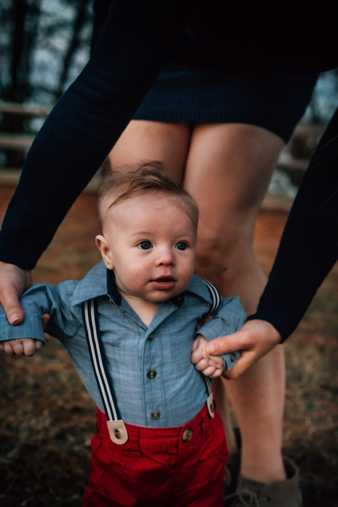 Hampton Roads family photographers capture little baby boy walking while wearing red pants, suspenders and a denim shirt as his mother holds his hands during their family photos