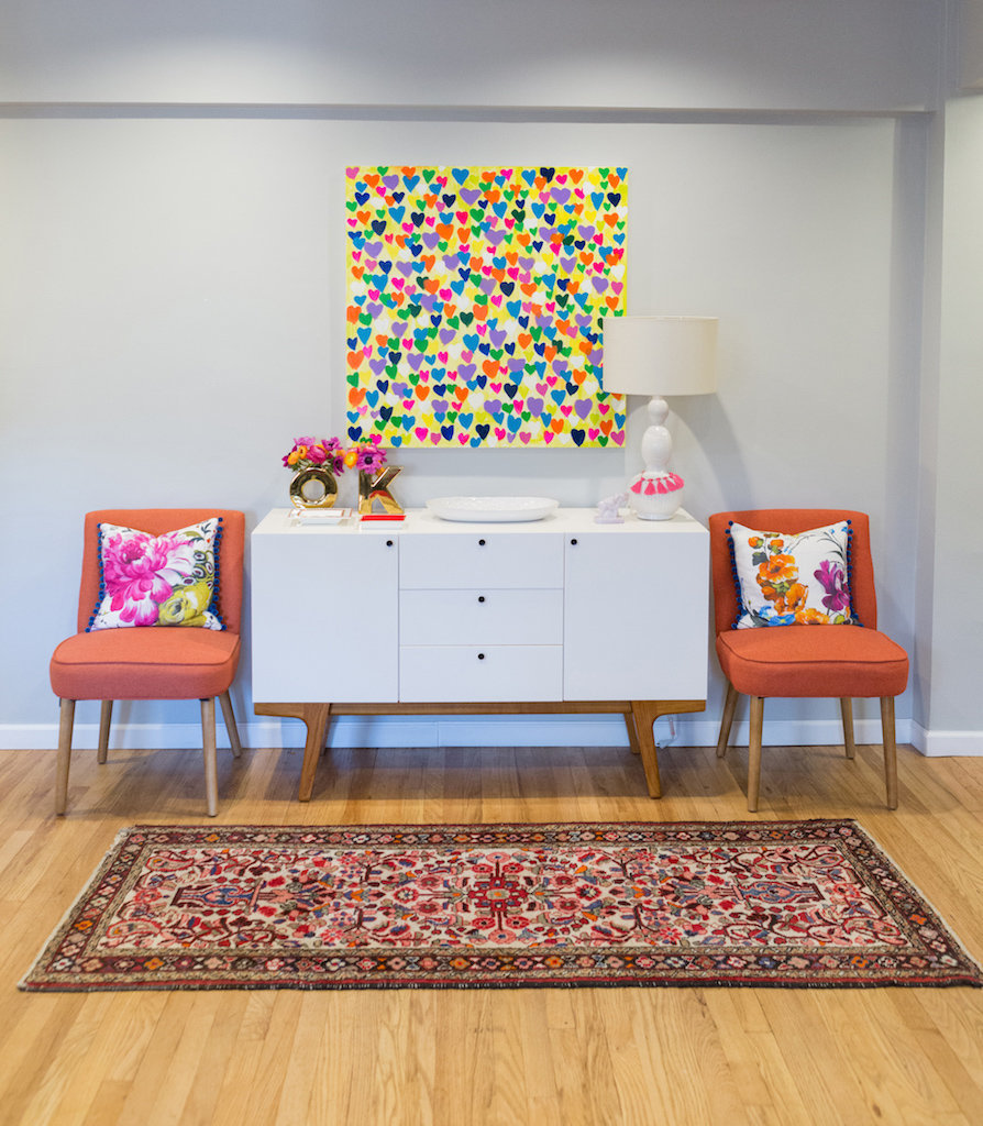 A modern white side table, colorful heart painting, and orange chairs.