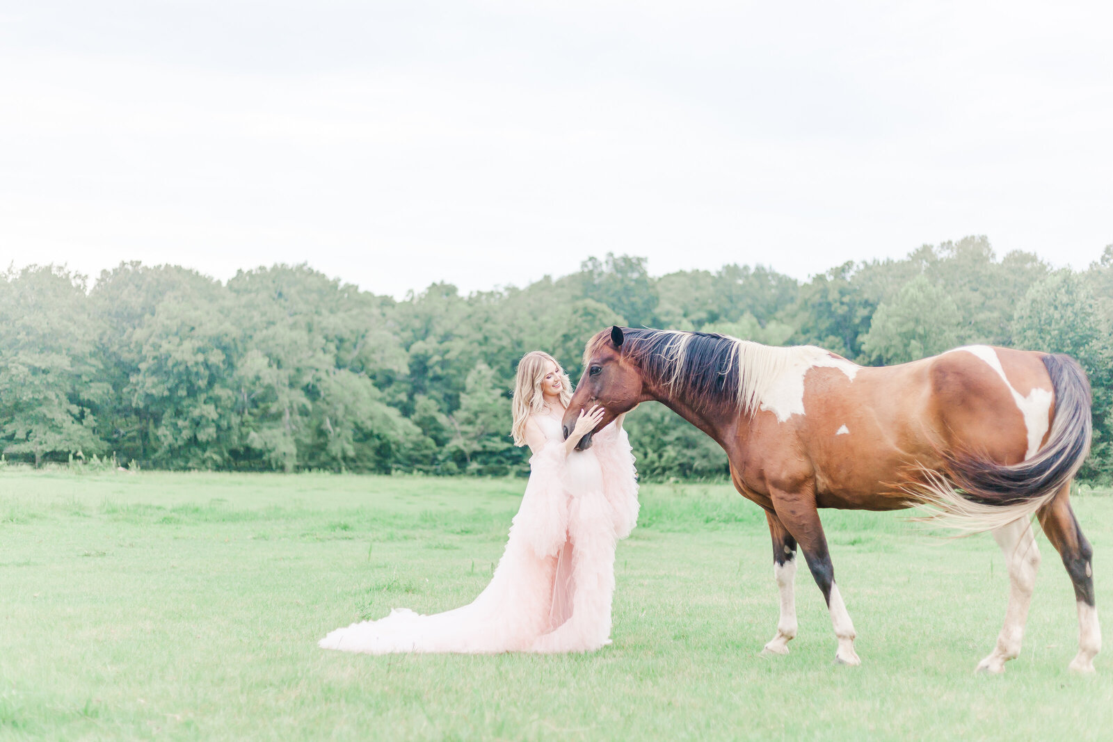 Pregnant woman wearing light pink dress petting horses nose in field during maternity session with 5U Photography in Birmingham, AL