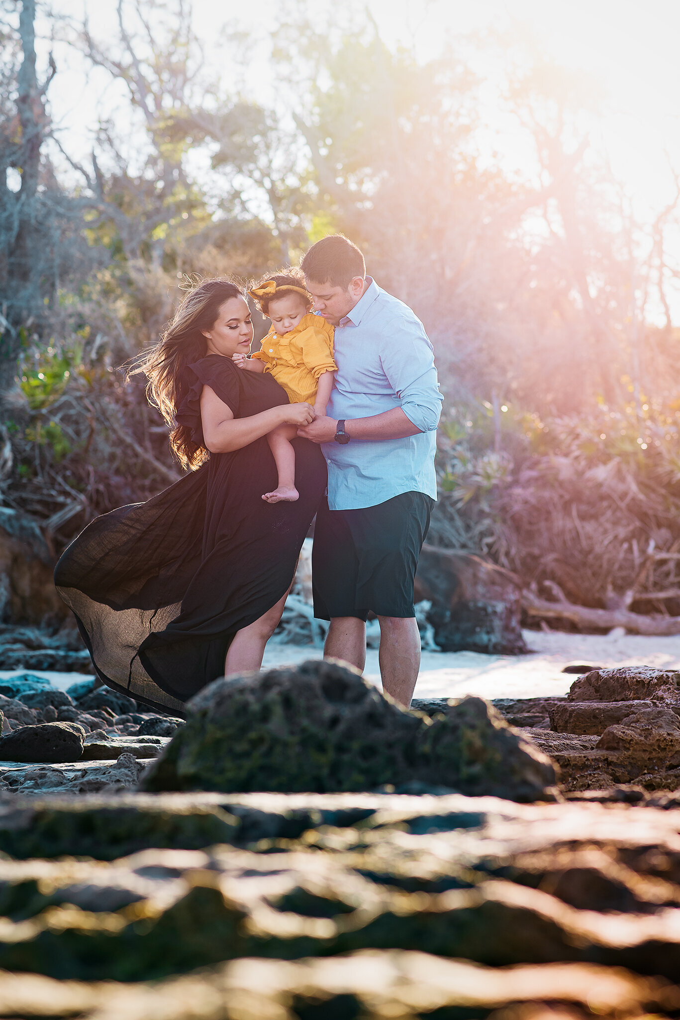 Pregnant mom in black dress holds daughter with husband at Black Rock Beach in FL.