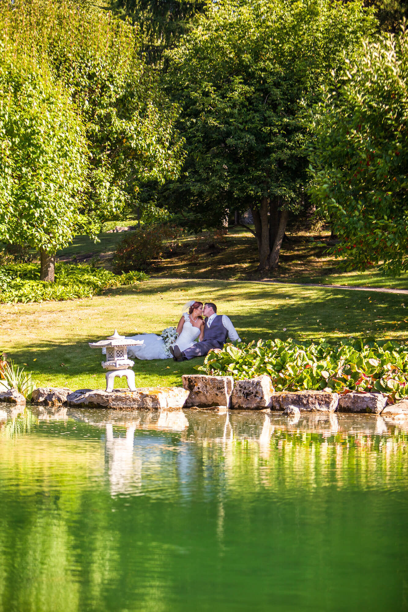 Bride and Groom sitting in the shade across the pond kissing