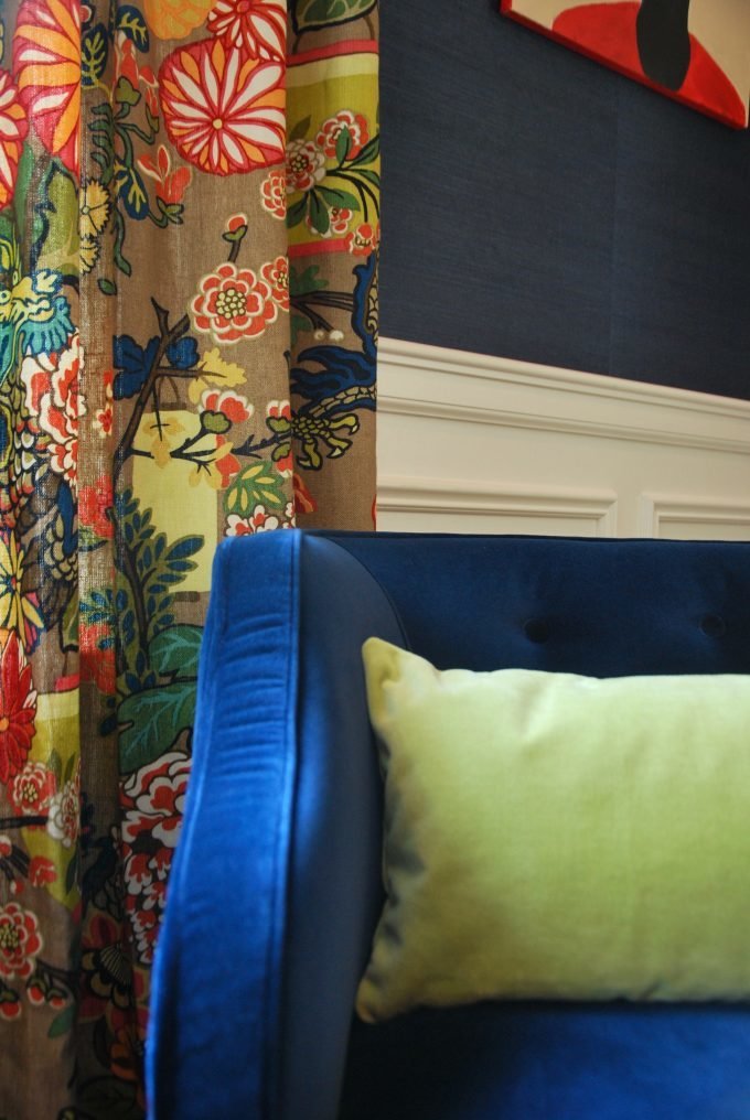 A blue velvet chair with lime green throw pillow in front of floral drapes.