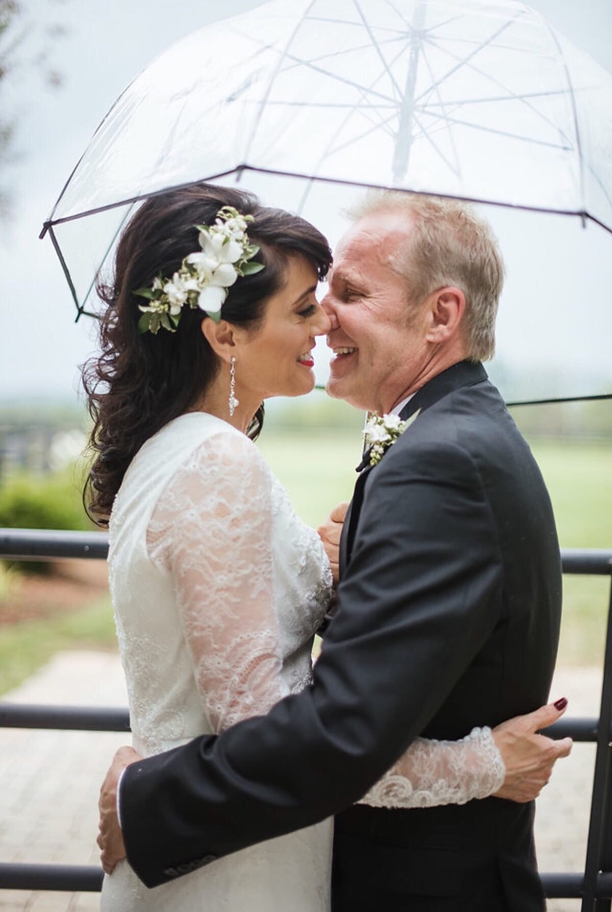 The bride, wearing a white suit with lace sleeves and the groom in a dark gray suit, embrace and kiss under a clear umbrella in the rain