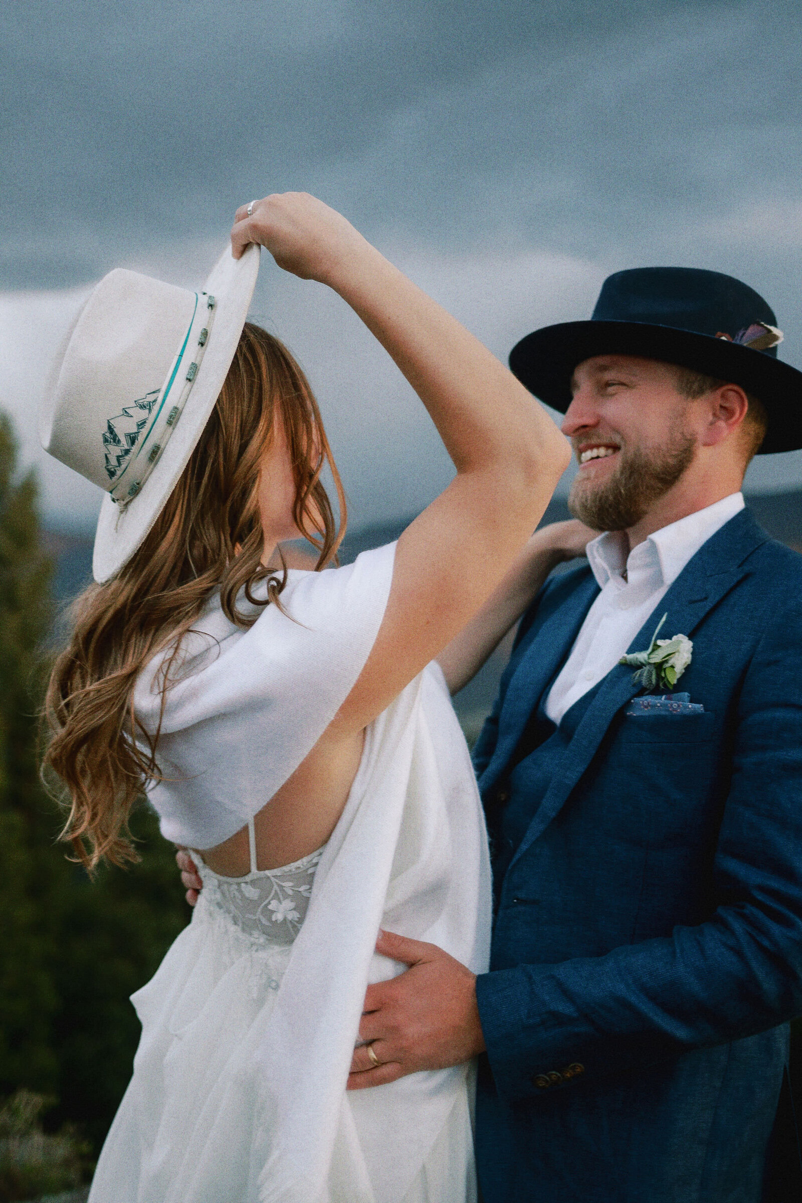 A groom with his hands around a bride's waist as she adjusts a hat.