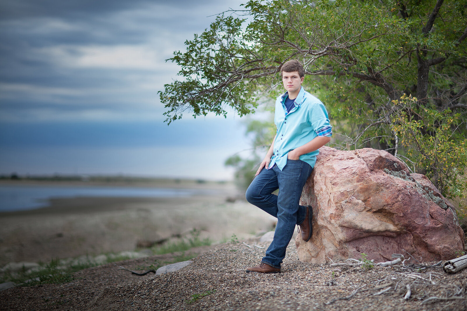 Graduation pictures. Picture of senior wearing teal shirt and blue jeans, leaning on rock next to lake. Senior Yearbook pictures in Billings Montana.
