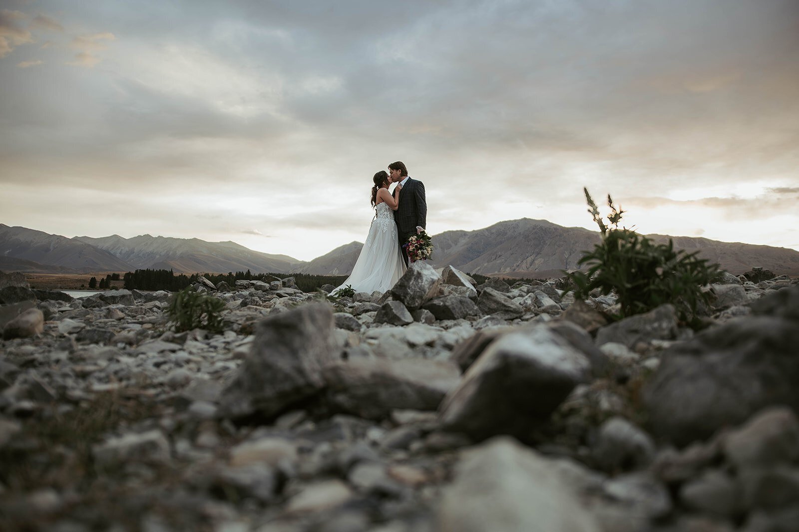 Bride and groom embrace on a rocky landscape with mountains and a dramatic sky in the background, capturing the spirit of an Oregon coast photography retreat for wild-hearted artists.