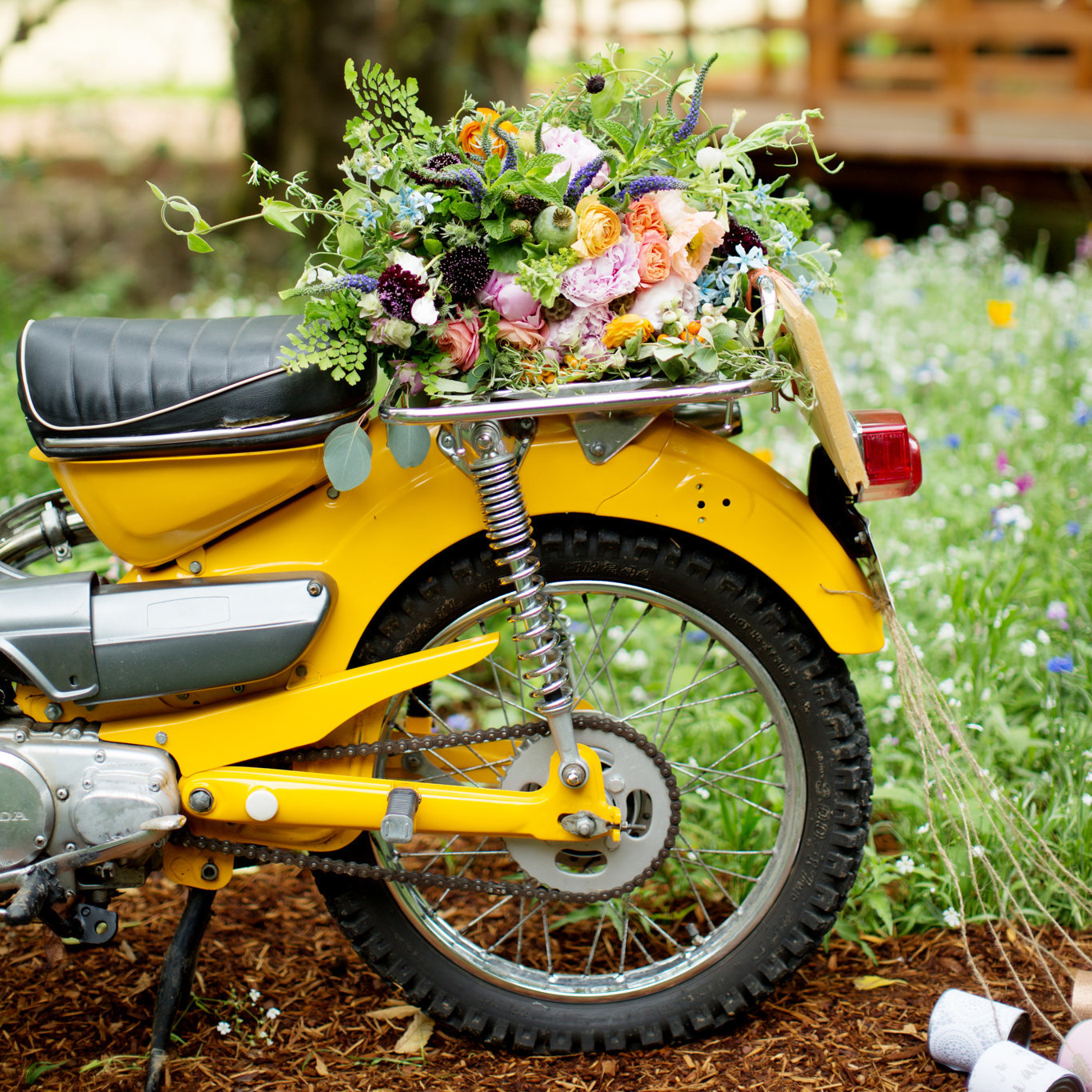a pretty wildflower bouquet sits atop a bright yellow motorcycle that has just married cans tied to the back