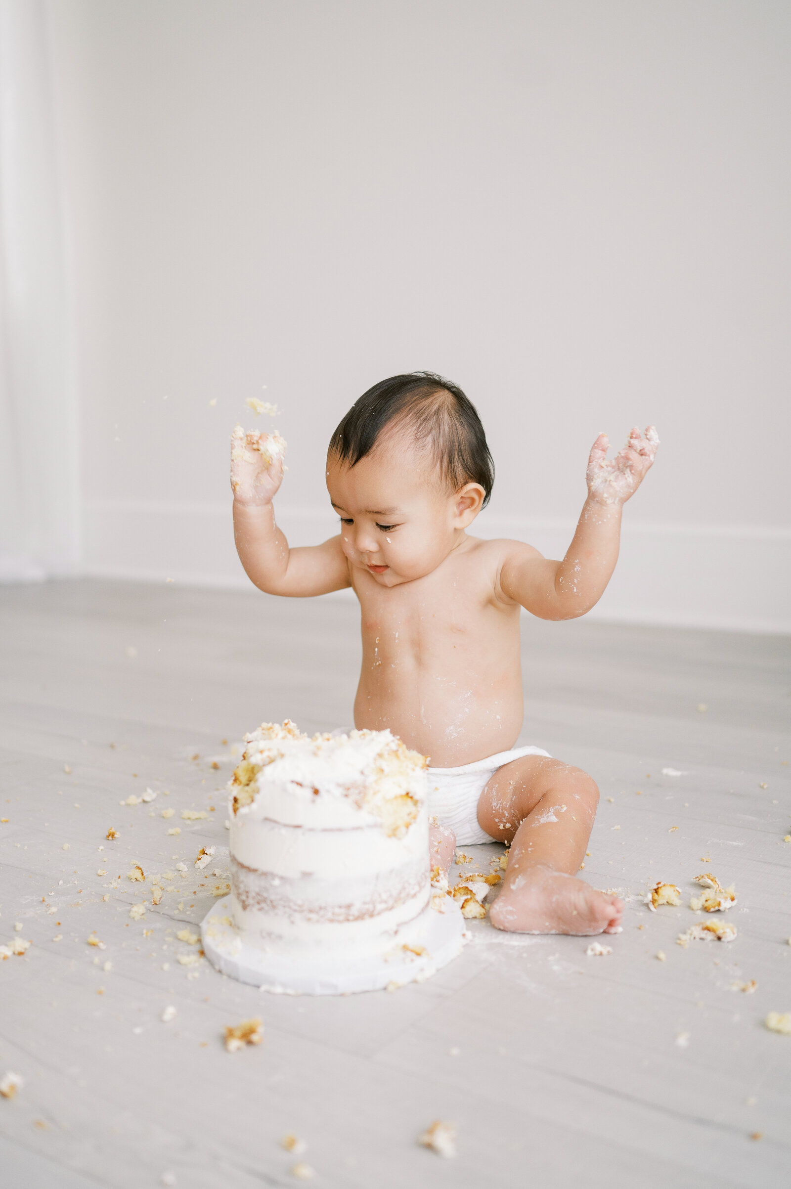 Baby boy in white diaper cover smashes white birthday cake during photo session with Worth Capturing Photography