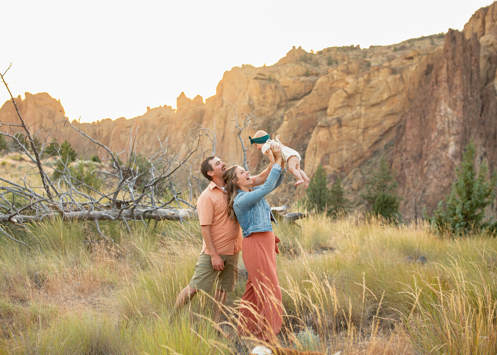 Family of three standing in field at sunset holding baby girl up in the air.