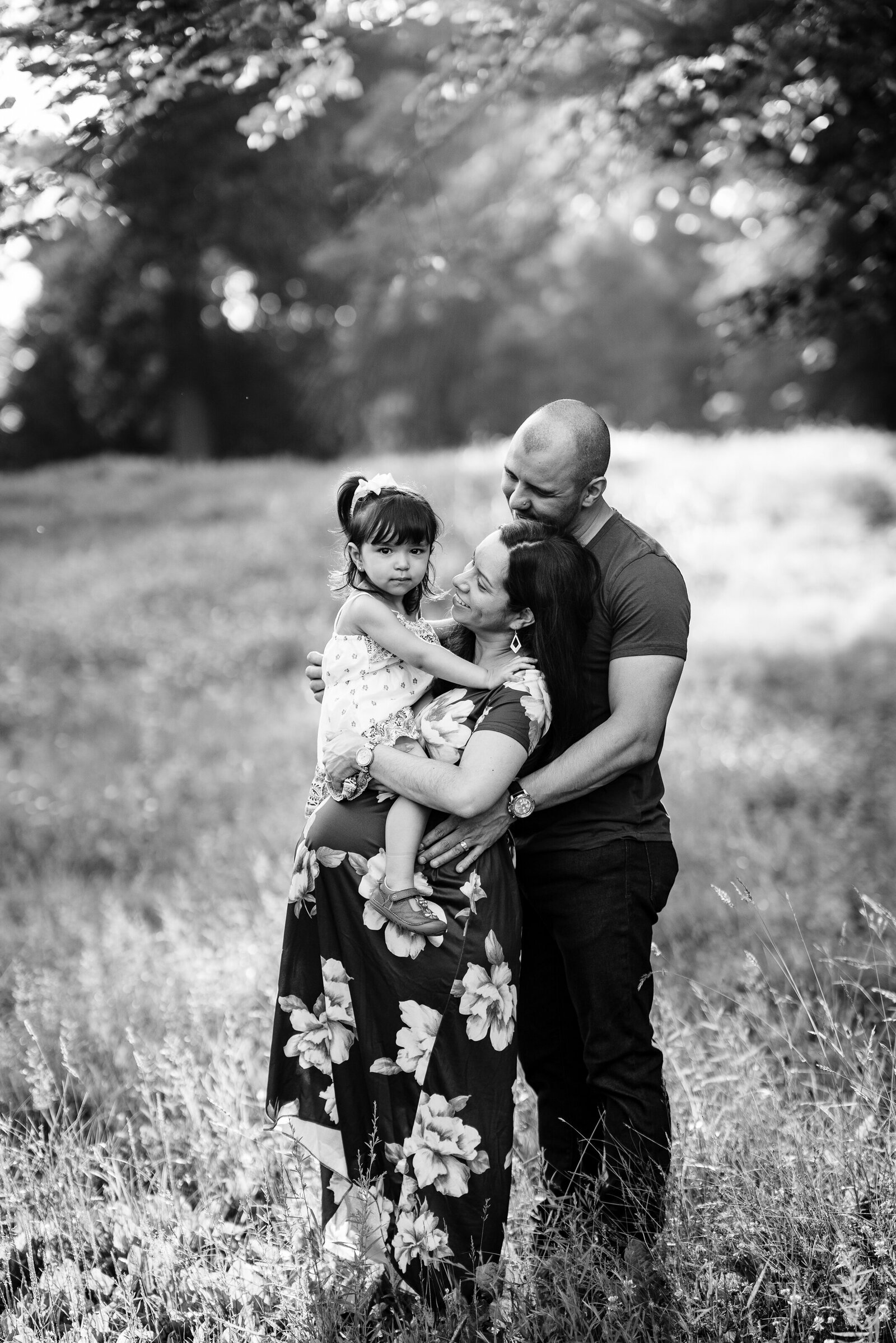 pregnant mother in a floral dress is hugged by father and toddler girl