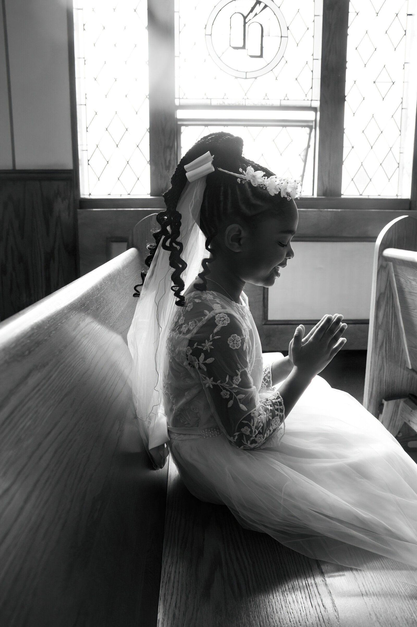 black and white photo of young black girl in First Communion veil and gown prays by a bright window New York First Communion photographer Milestone Images