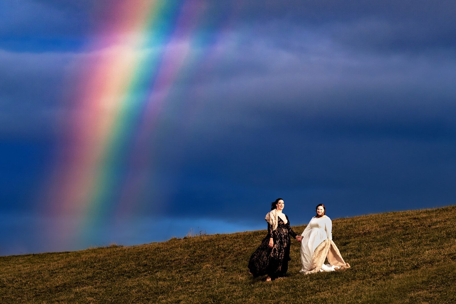 creative wedding photography of a same-sex couple standing in a field with a rainbow in the background