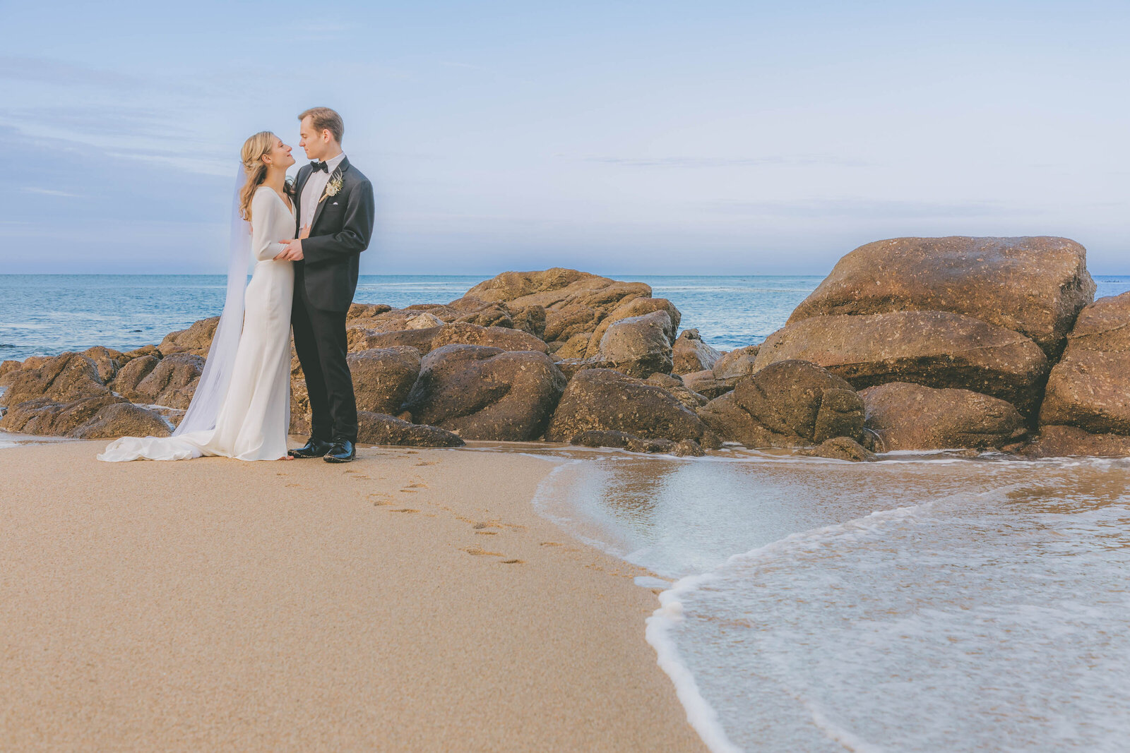 A bride and groom are towards the left of the photo as the ocean's waves flow towards the right during their beach wedding.