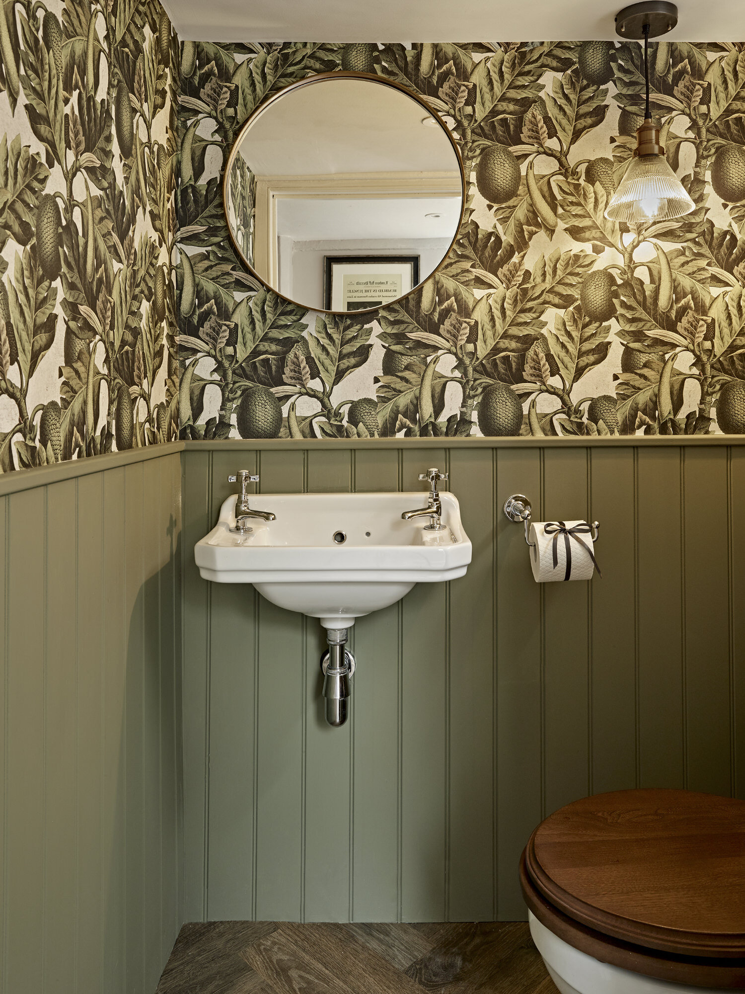 olive green wall with tropical leaf batten wall paper in bathroom with sink and round mirror