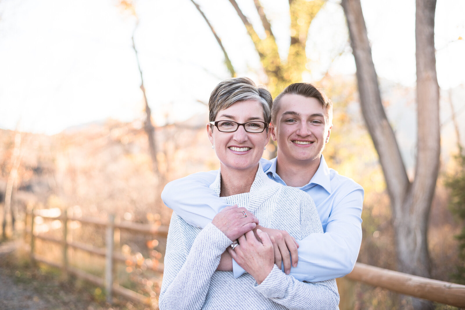 denver family photographers captures teenage child hugging his mom around the shoulders as he stands behind her on a beautiful fall path in denver