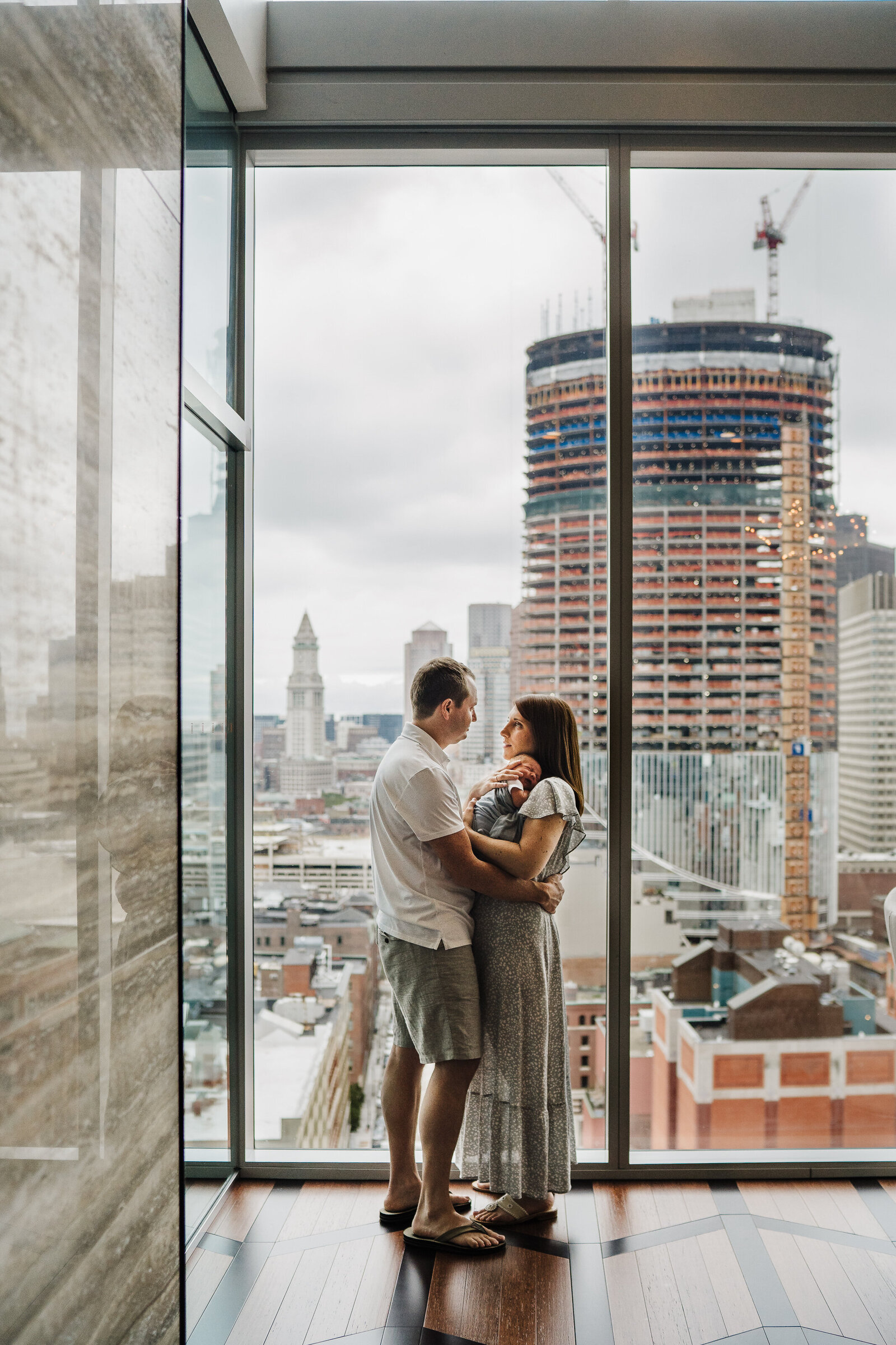 parents holding new baby stand in front of huge windows overlooking boston cityscape
