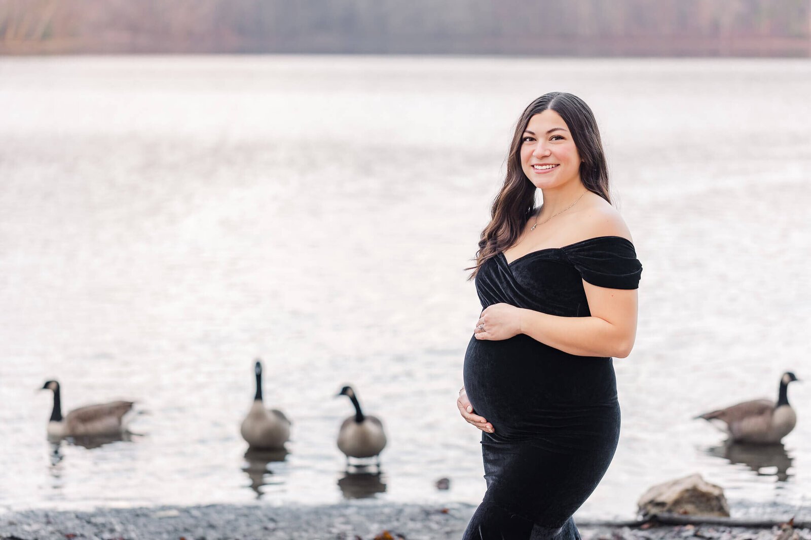 A pregnant woman in a black dress posing in front of a Burke Lake full of geese.